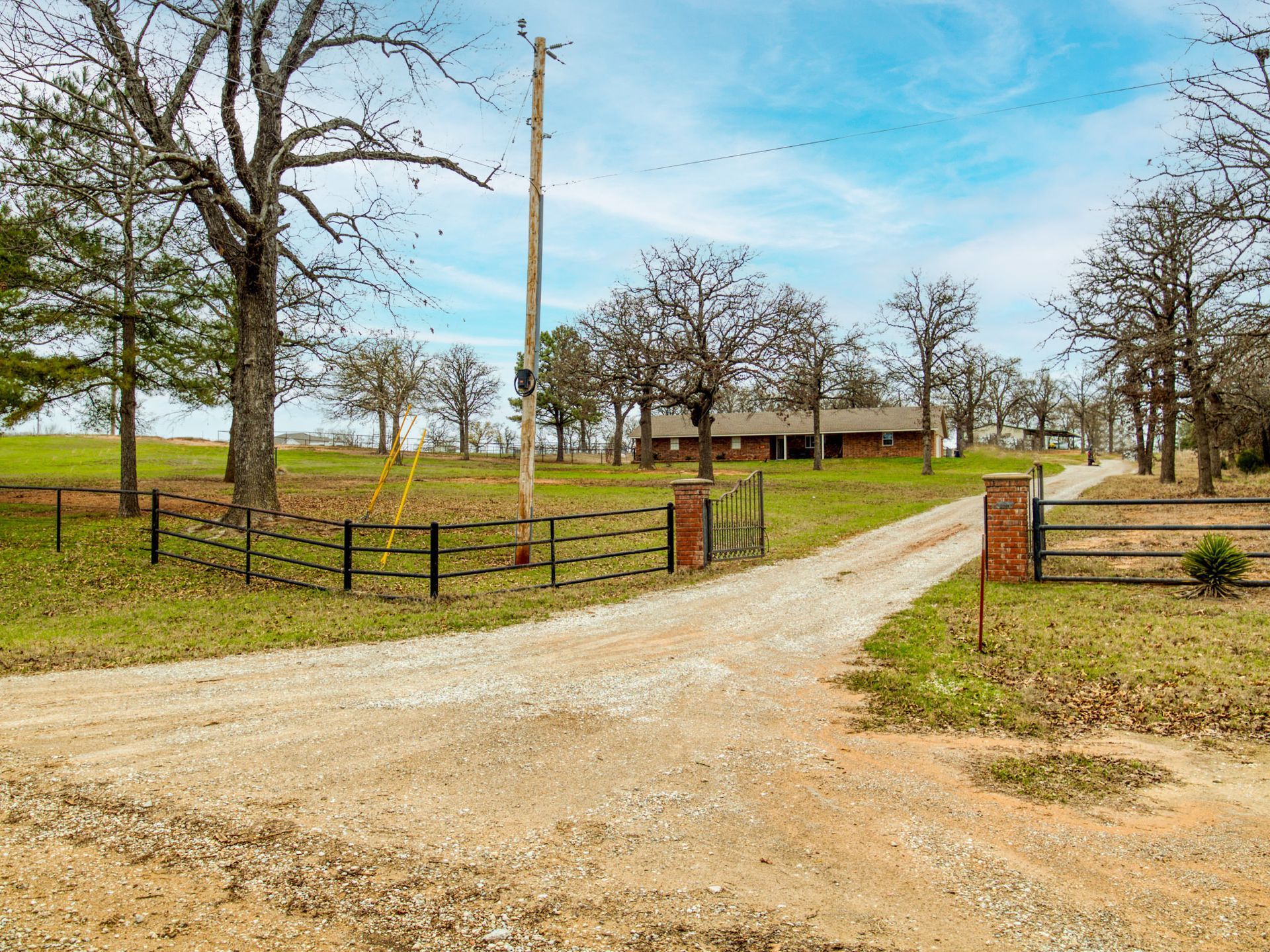 Gravel driveway leads to a single-story brick house, with a fence and trees surrounding the property.