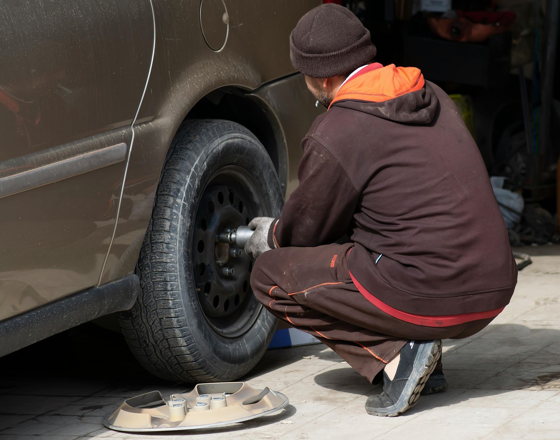 A person wearing a beanie and a brown hoodie squats to tighten the lug nuts on a car tire with a power tool.