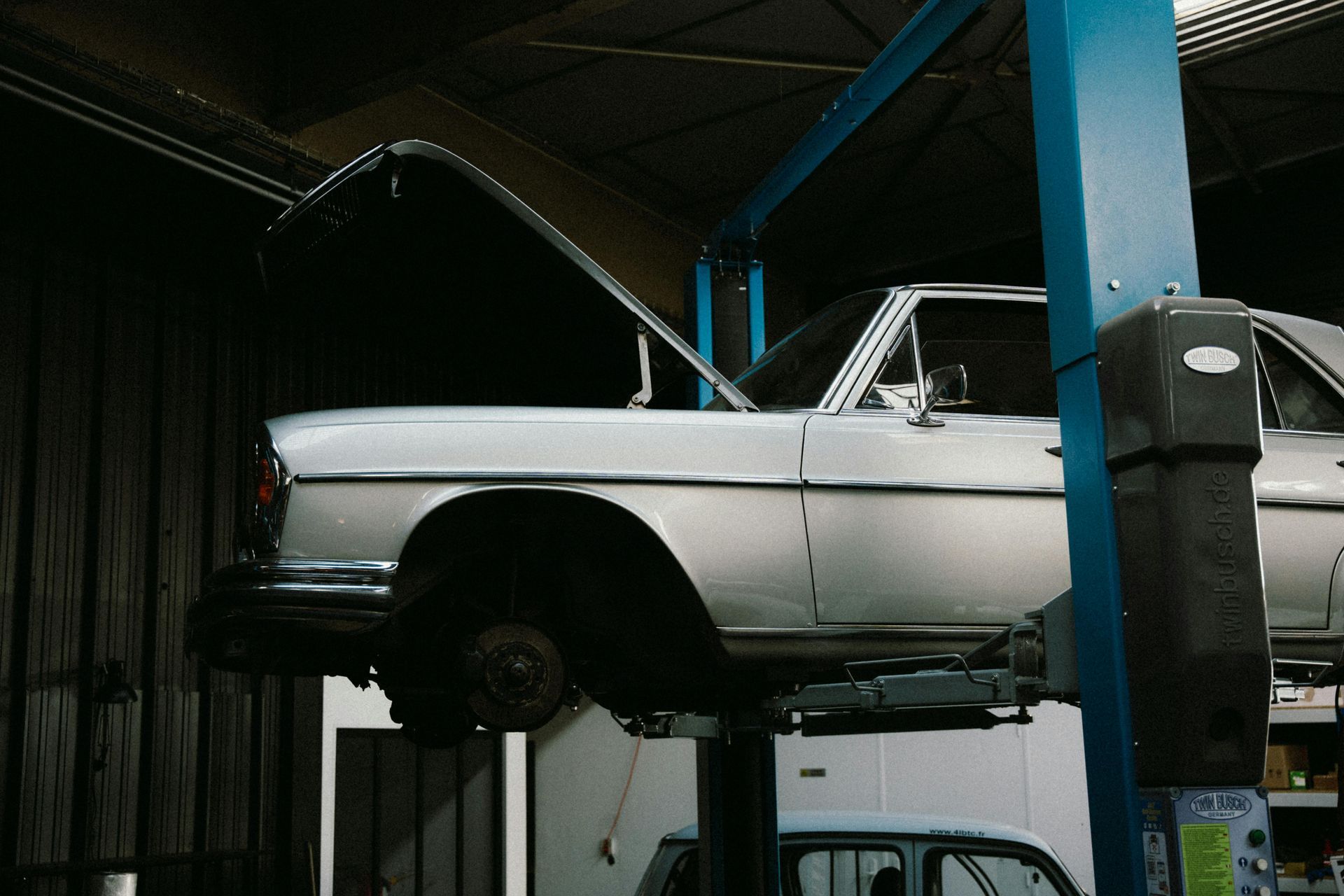A silver vintage car with its hood open, raised on a blue hydraulic lift inside an auto repair shop.