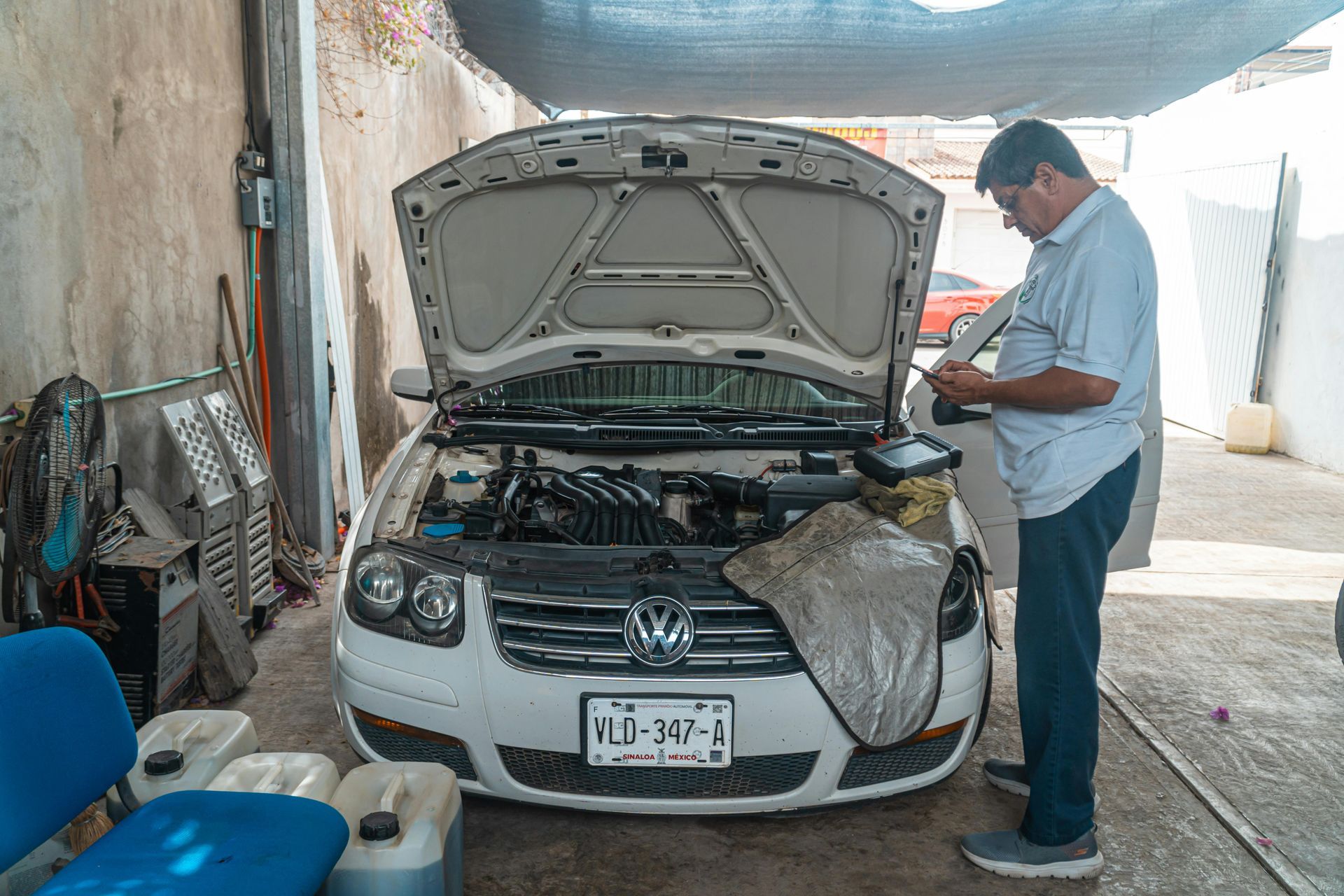 A person in a light blue shirt works on the engine of a white Volkswagen car with its hood open in a repair shop.