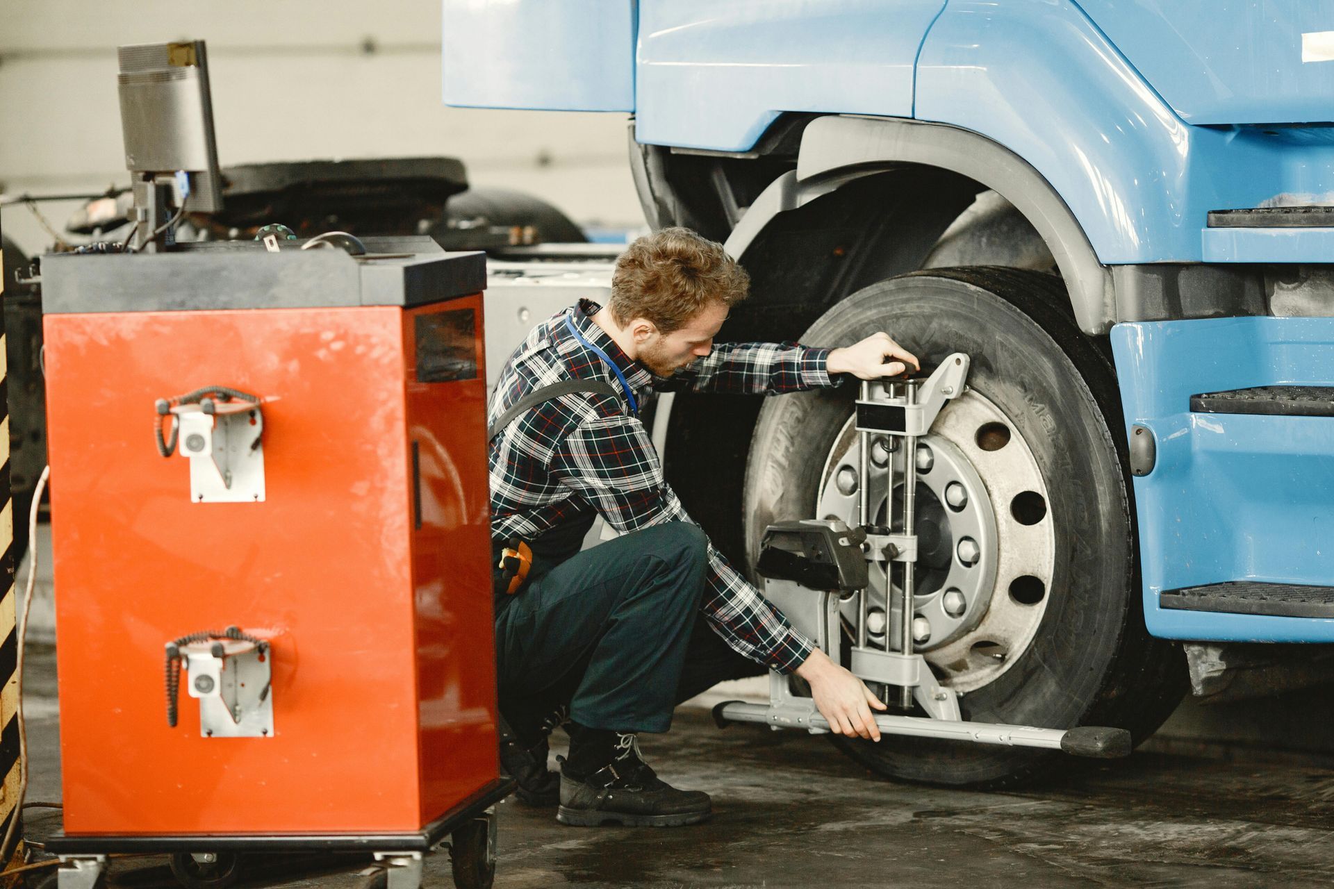 A mechanic in a plaid shirt attaches a diagnostic alignment tool to the wheel of a blue truck in a workshop.