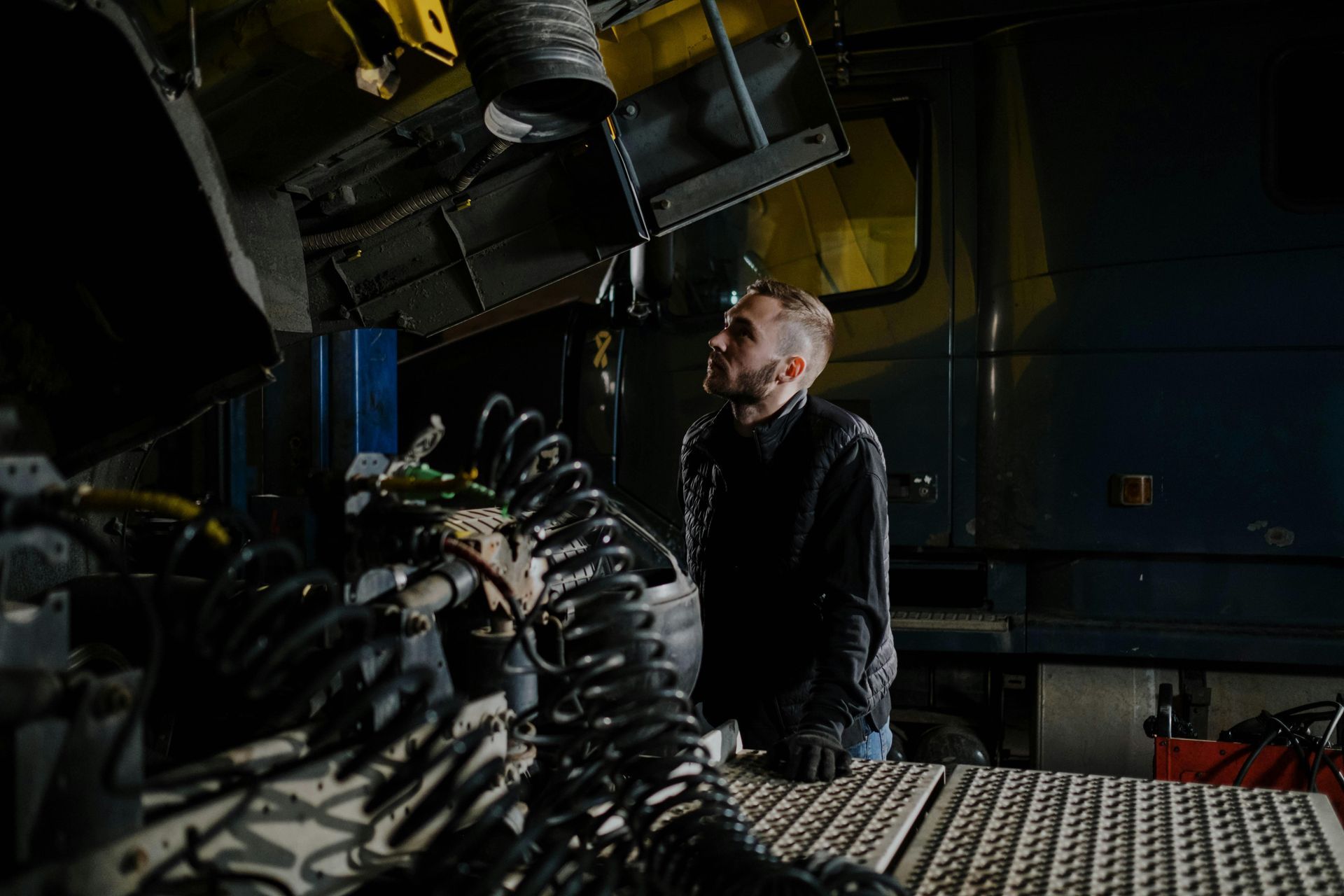 A person in a dark jacket inspects the engine of a truck in a workshop with a raised hood.