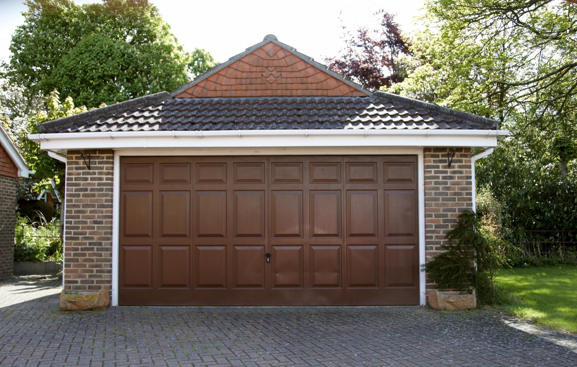 Brown garage door on brick building with a tiled roof, set on a patterned driveway, surrounded by greenery.