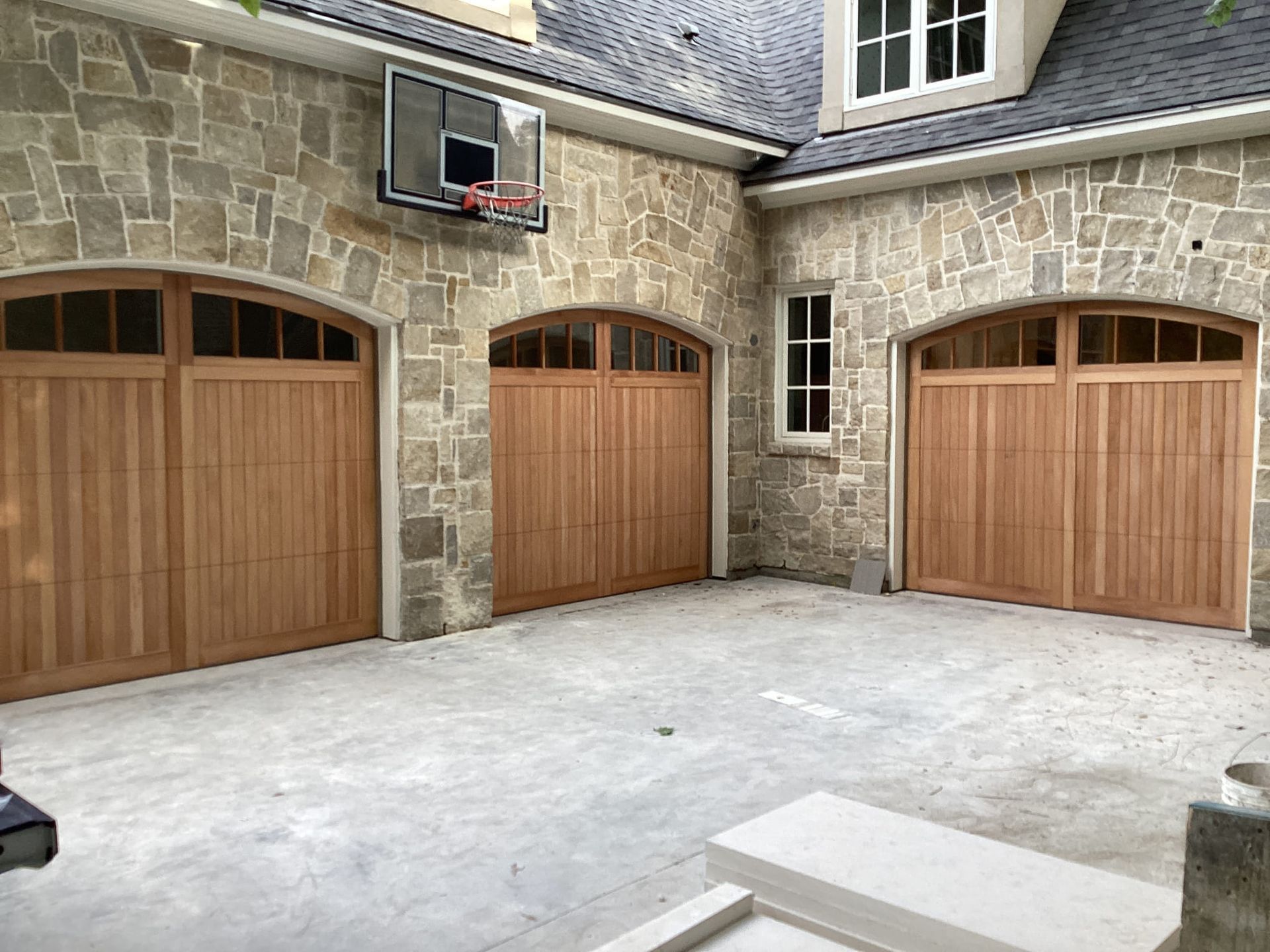 A stone house exterior with three arched wooden garage doors and a basketball hoop mounted above the left door.
