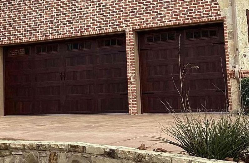 Two dark brown garage doors on a brick building with a concrete driveway and landscaping.