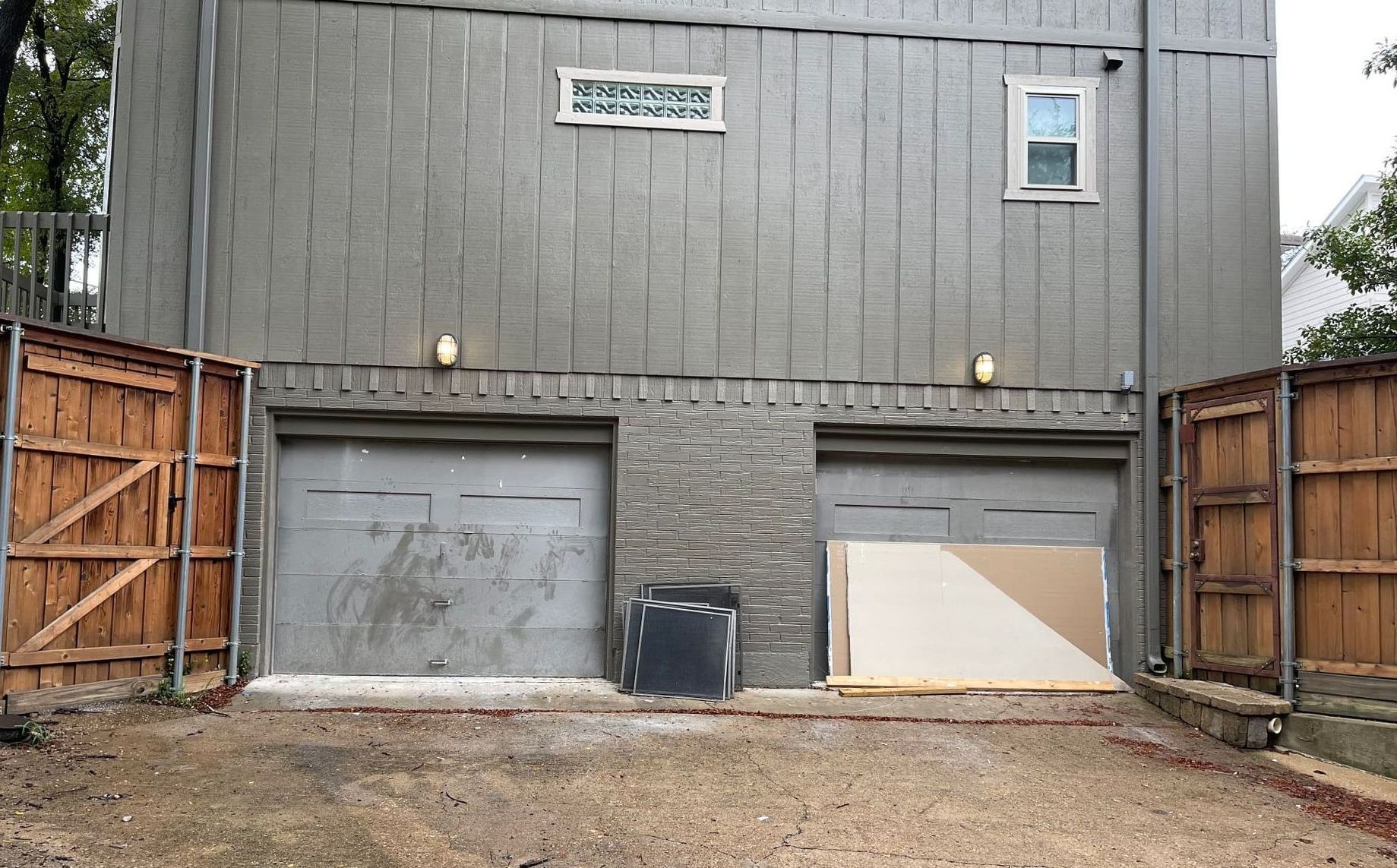 Two gray garage doors under a gray building, flanked by wooden fences. Gravel driveway.
