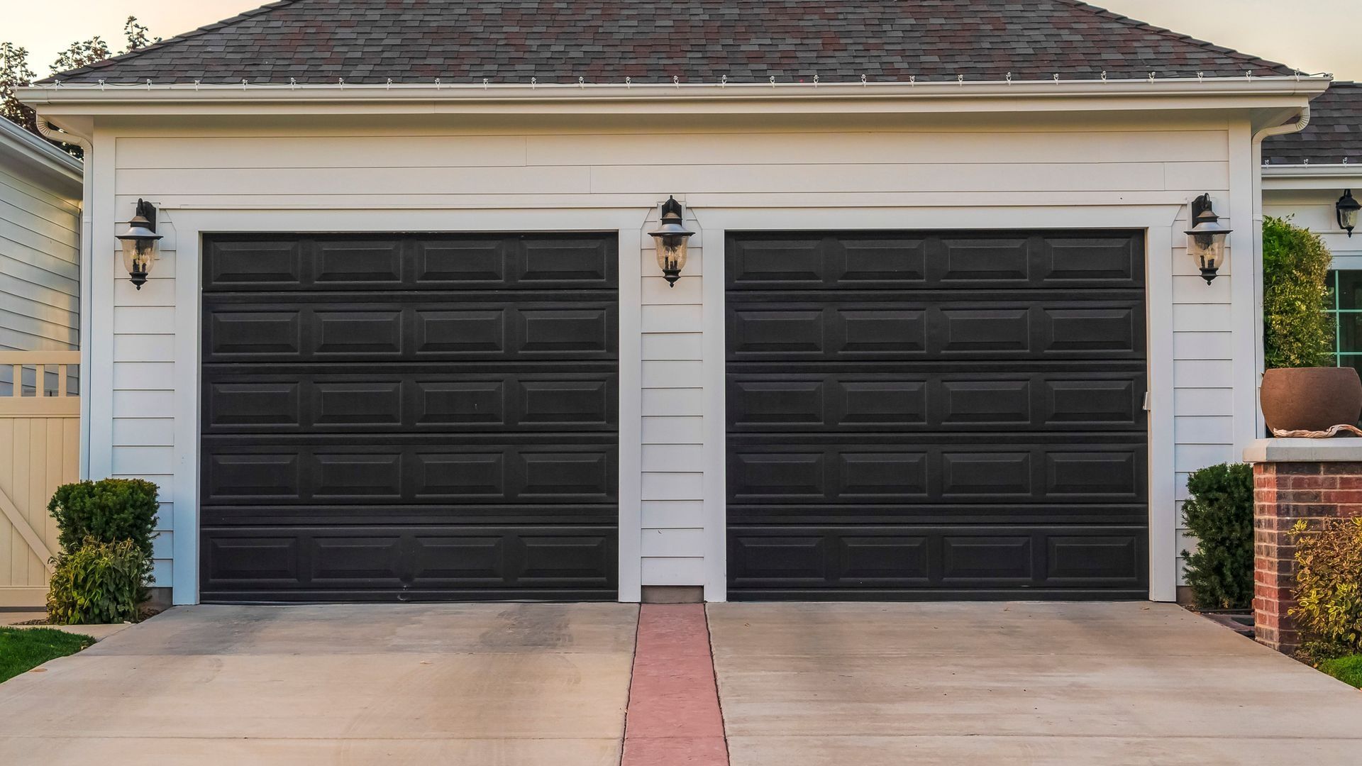 Two black garage doors on a white house, with a concrete driveway and brick accents.