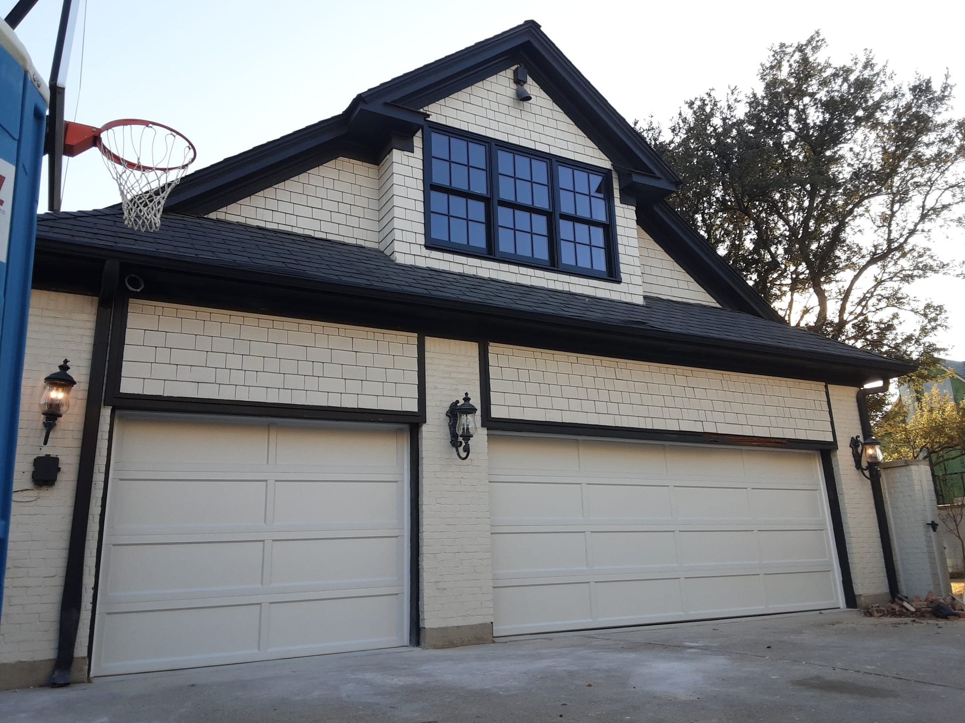 Two-car garage with black trim, tan brick, and a small loft with dark windows. A basketball hoop is attached.