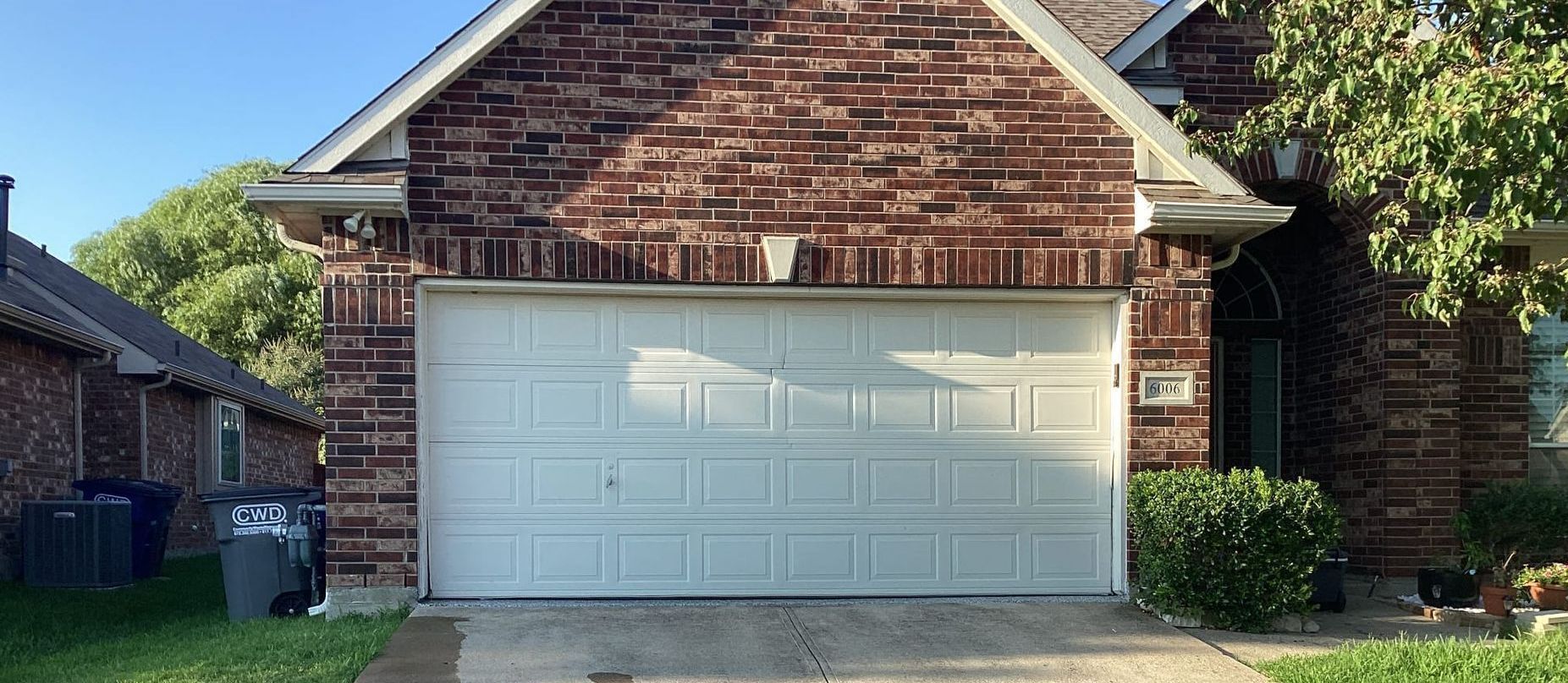 A brick house with a white garage door, trees, and trash cans.