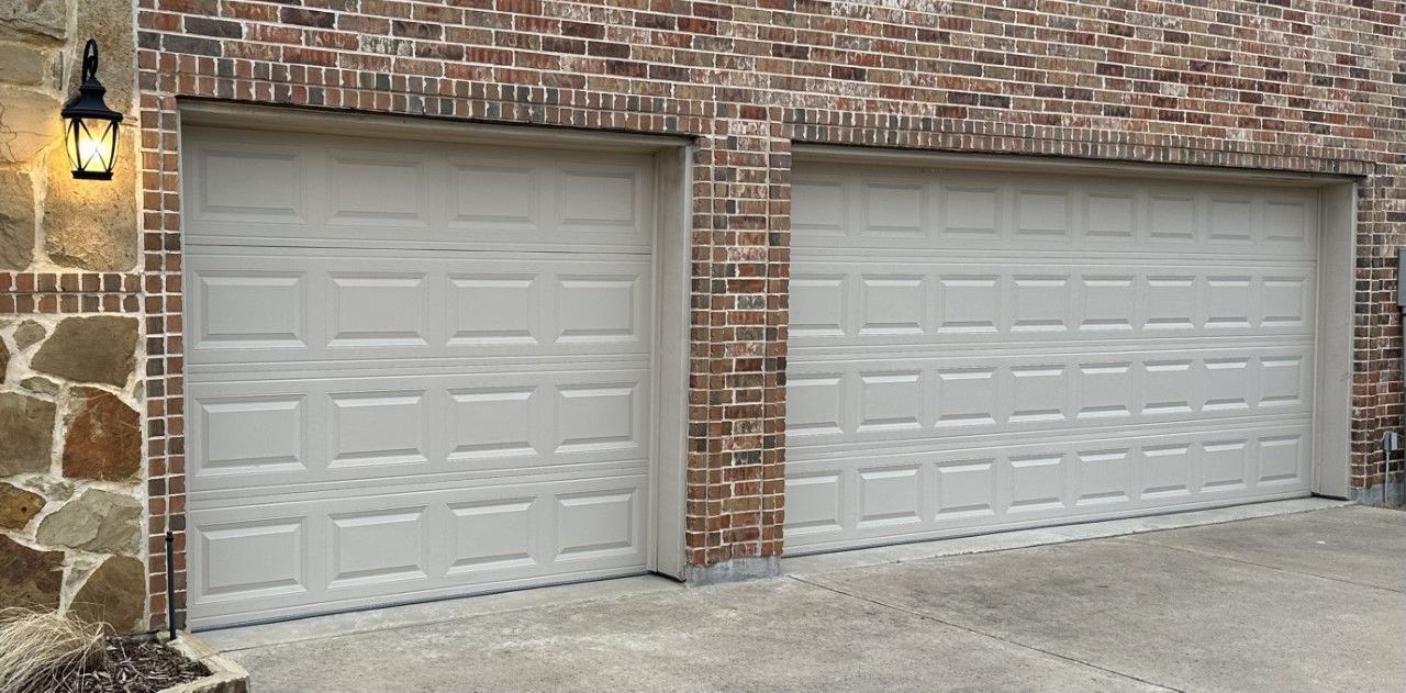 Two beige garage doors framed by brick and stone, a lit lamp on the left.
