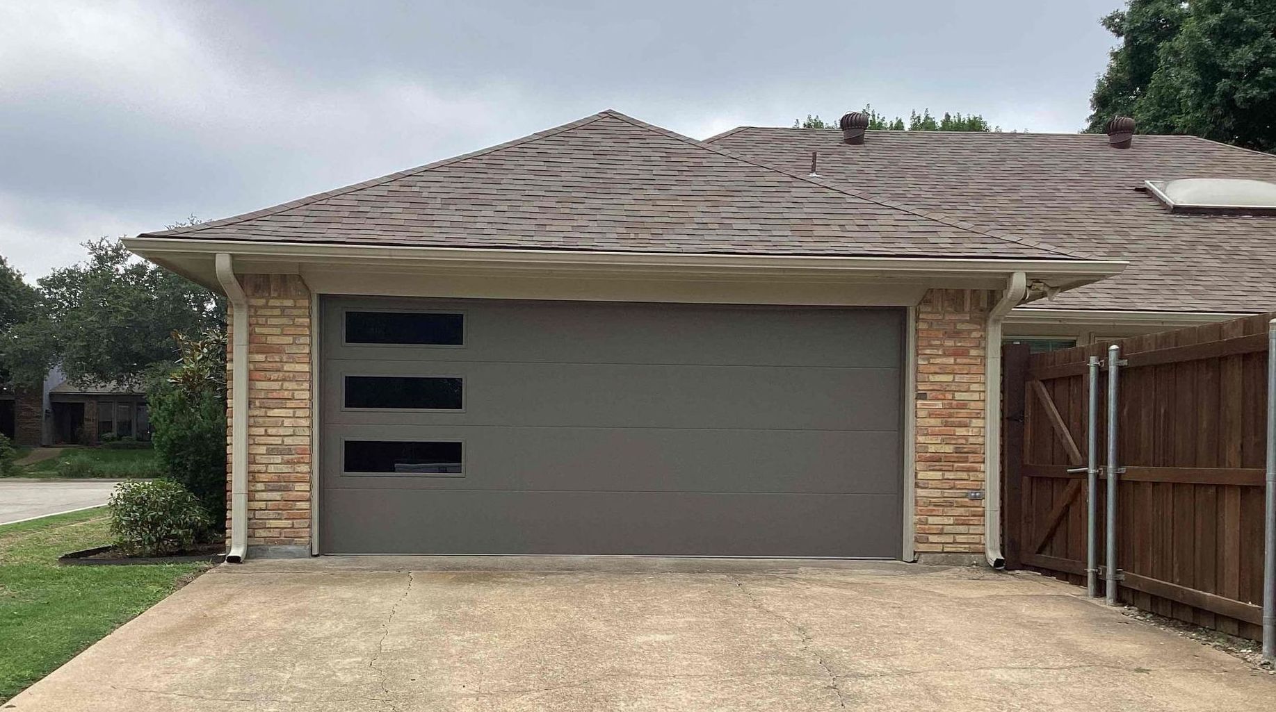 Gray garage door with three horizontal windows, tan stone facade, and brown roof.