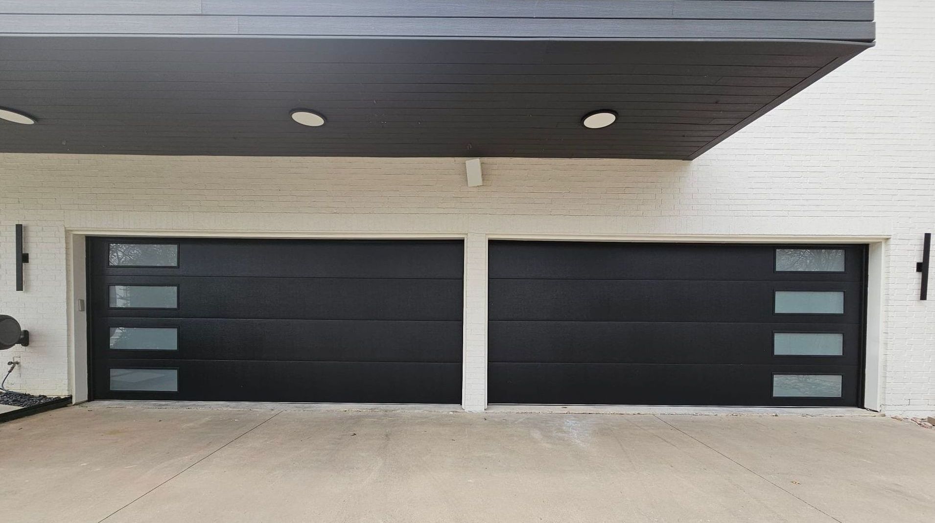 Two black garage doors with glass panels under a white brick and dark wooden overhang.