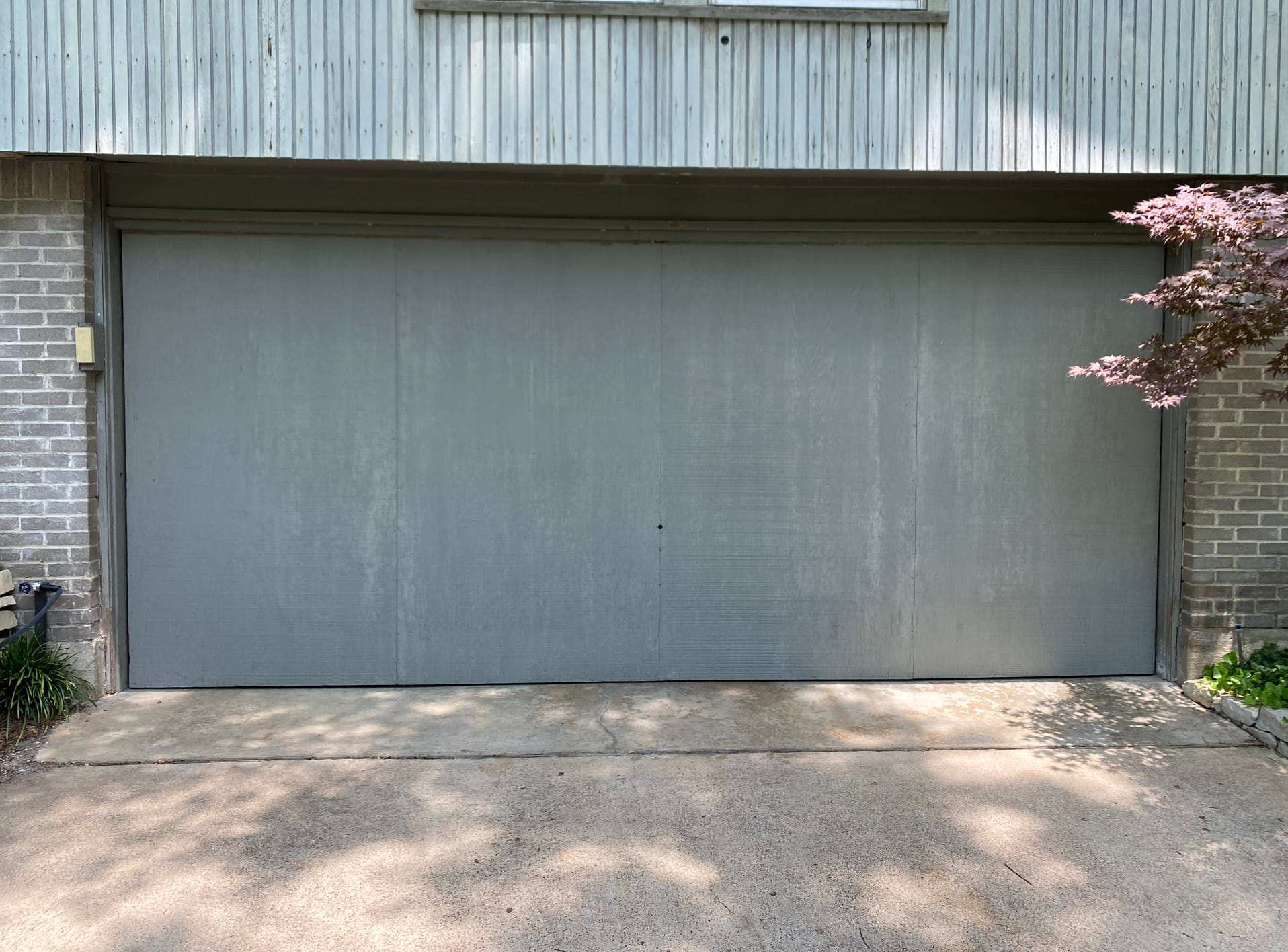 Gray garage door on a concrete driveway, flanked by brick and a small tree.
