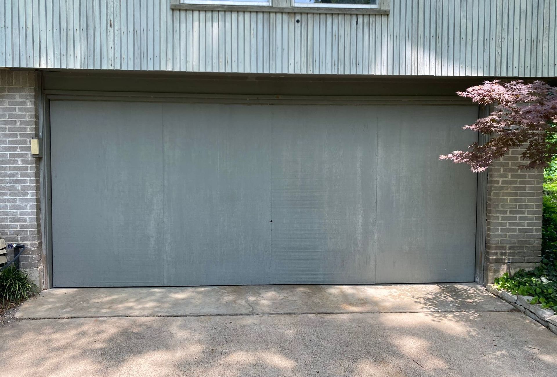 Gray garage door on a concrete driveway, flanked by brick and a small tree.