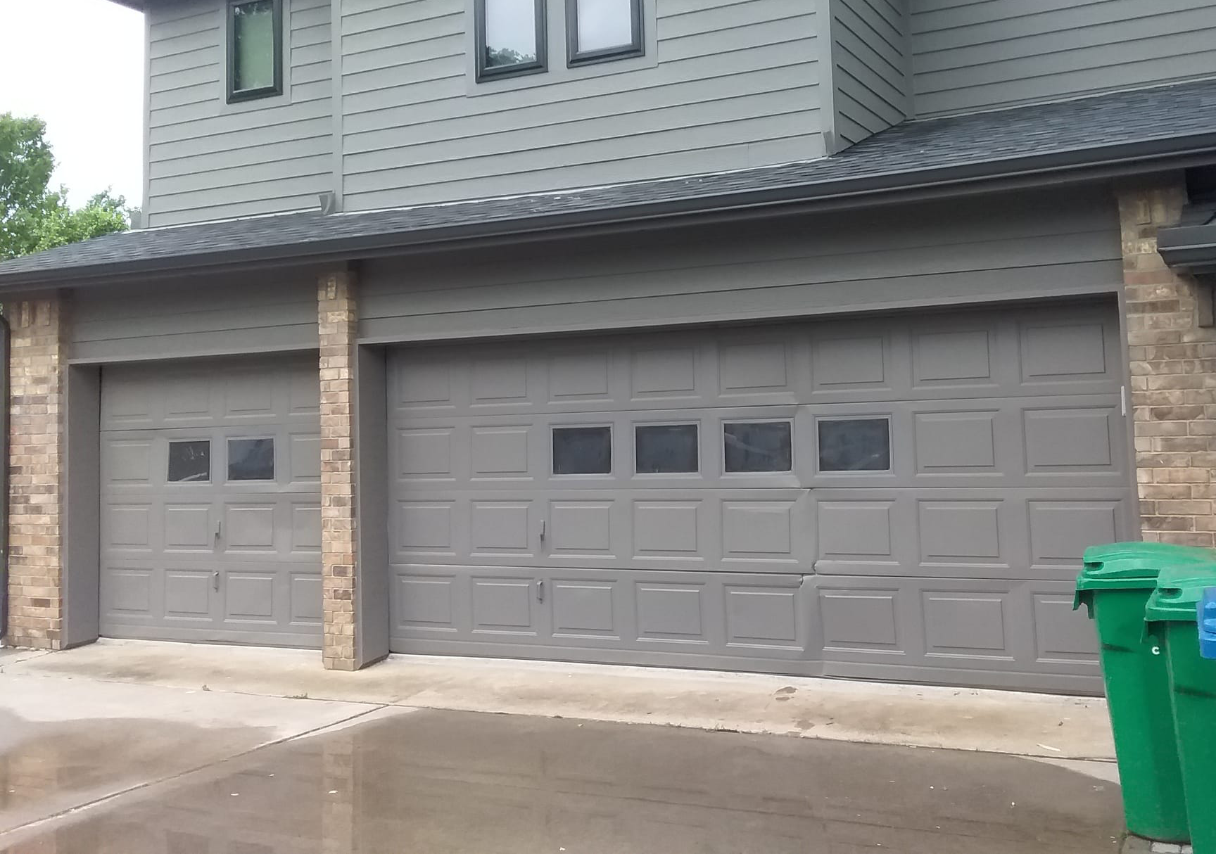 Gray garage doors with windows, flanked by brick columns and trash cans.
