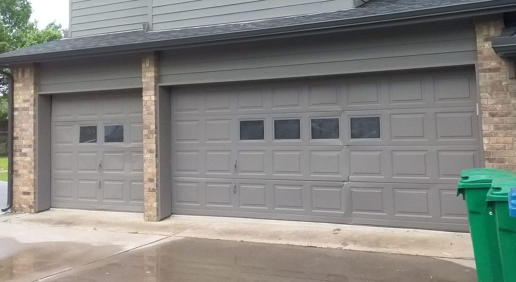 Gray garage doors with windows, flanked by brick columns and trash cans.