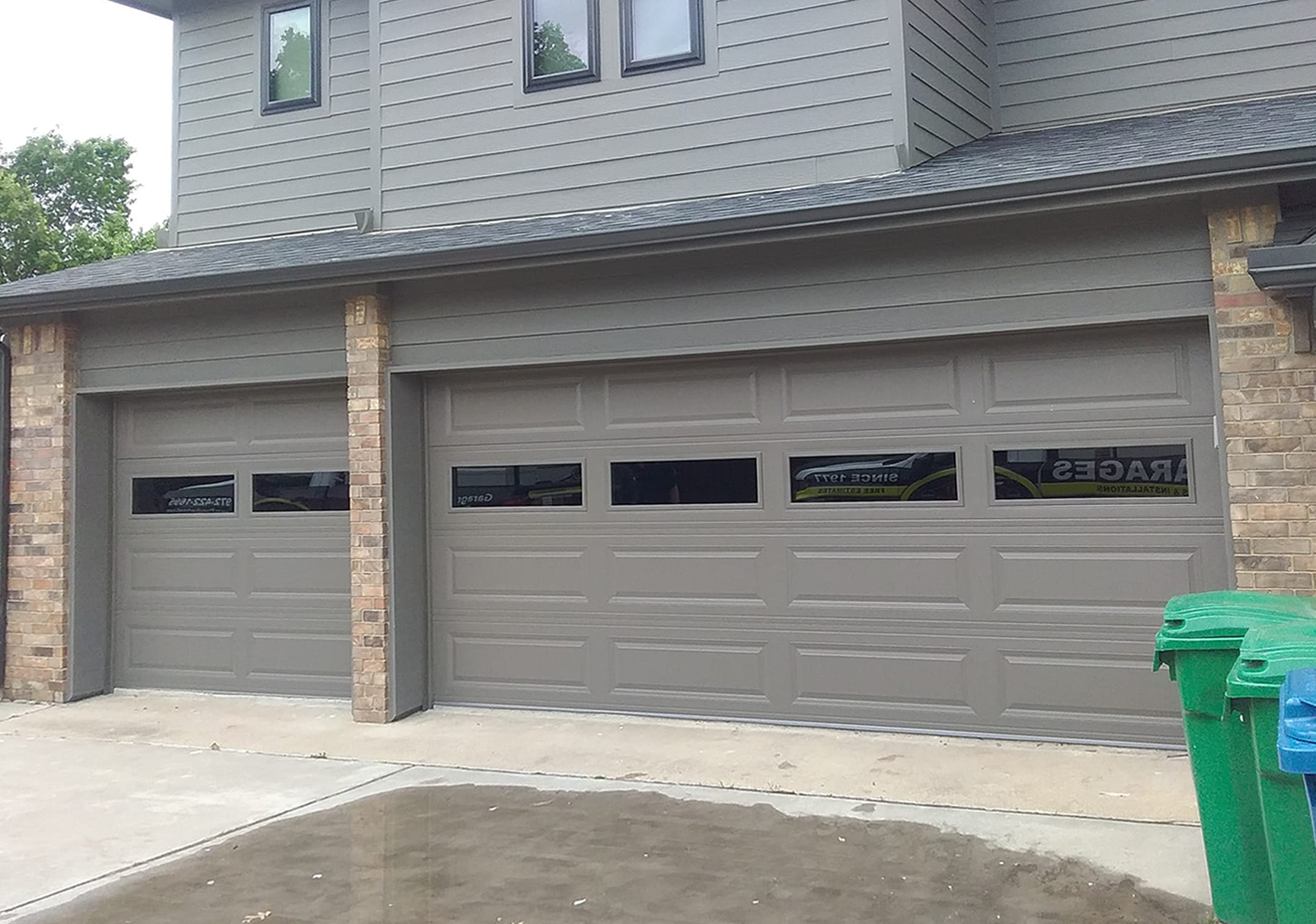 Two gray garage doors with window panels, in a brick building. Green and blue trash bins sit to the right.