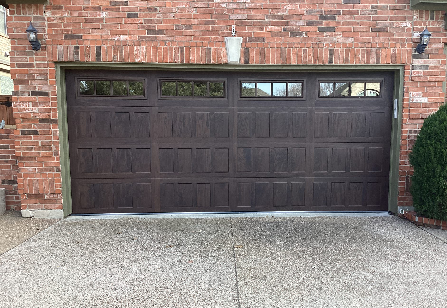 Brown garage door with windows, on a brick house. Concrete driveway with a bush to the right.