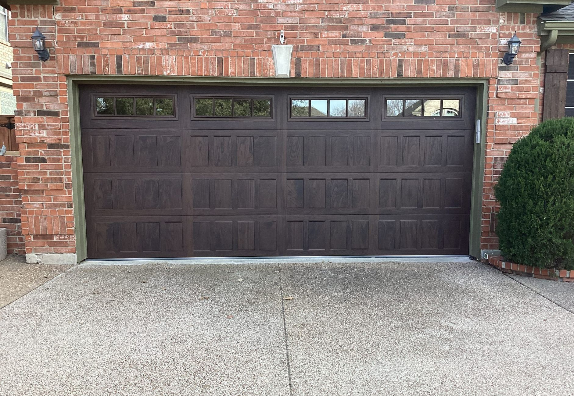 Brown garage door with windows, on a brick house. Concrete driveway with a bush to the right.
