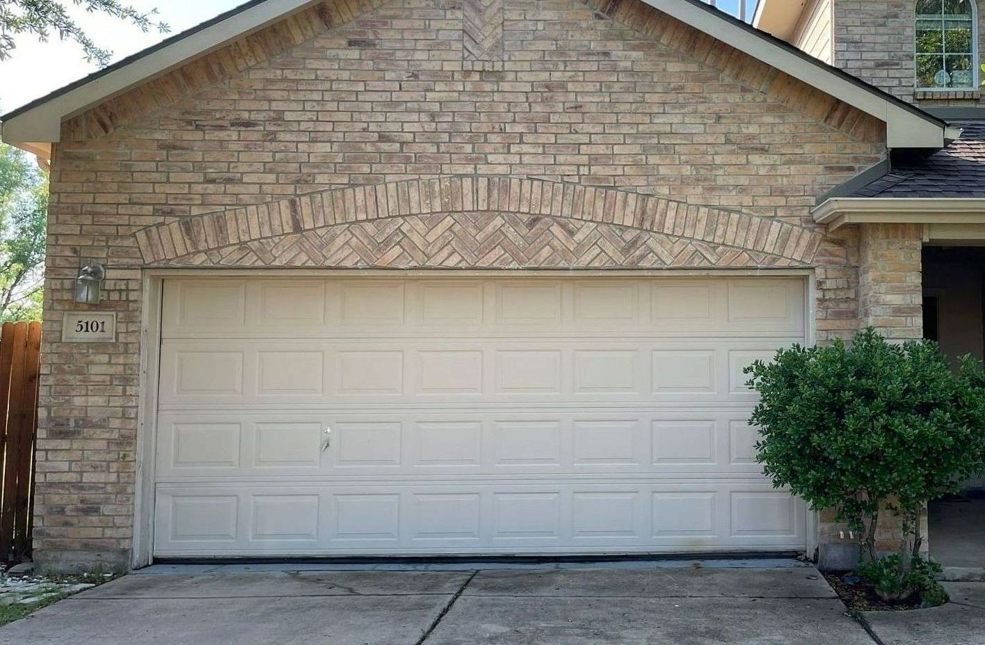 Beige garage door on a brick house with an arched brick design above the door. A small bush is to the right.