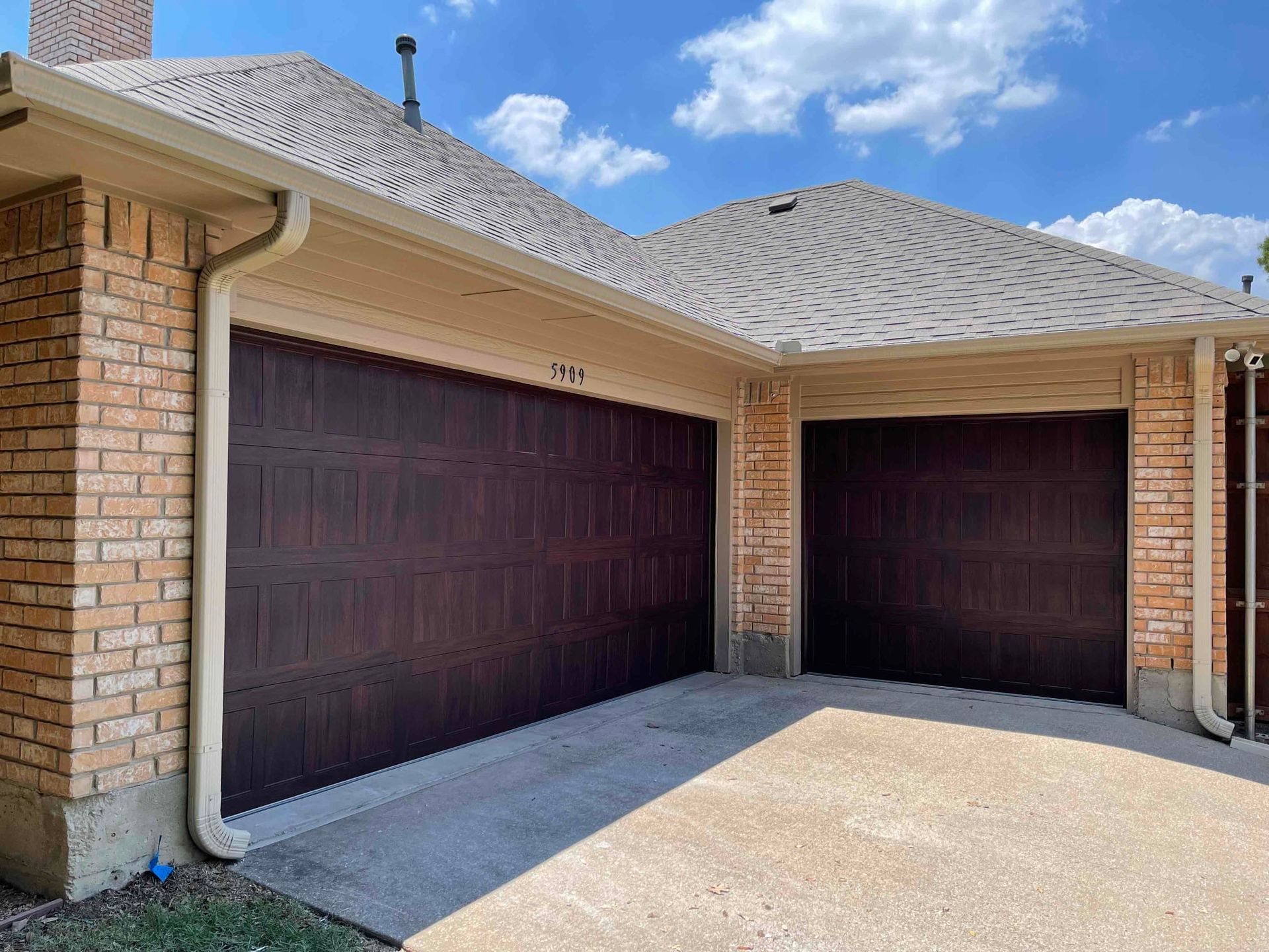 A tan brick house with two dark brown, paneled garage doors under a shingled roof on a sunny day.