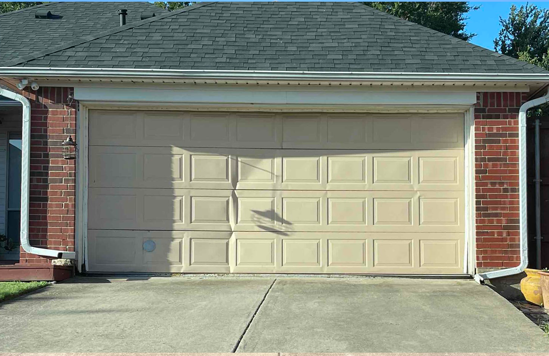 Tan garage door with shadow, brick siding, concrete driveway, and green grass.