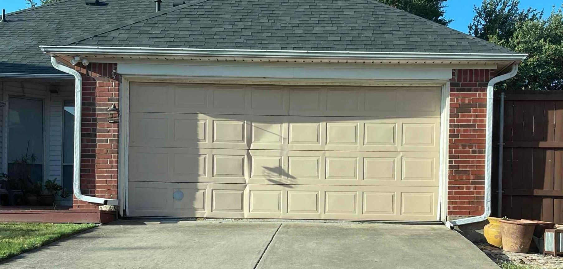 Tan garage door with shadow, brick siding, concrete driveway, and green grass.