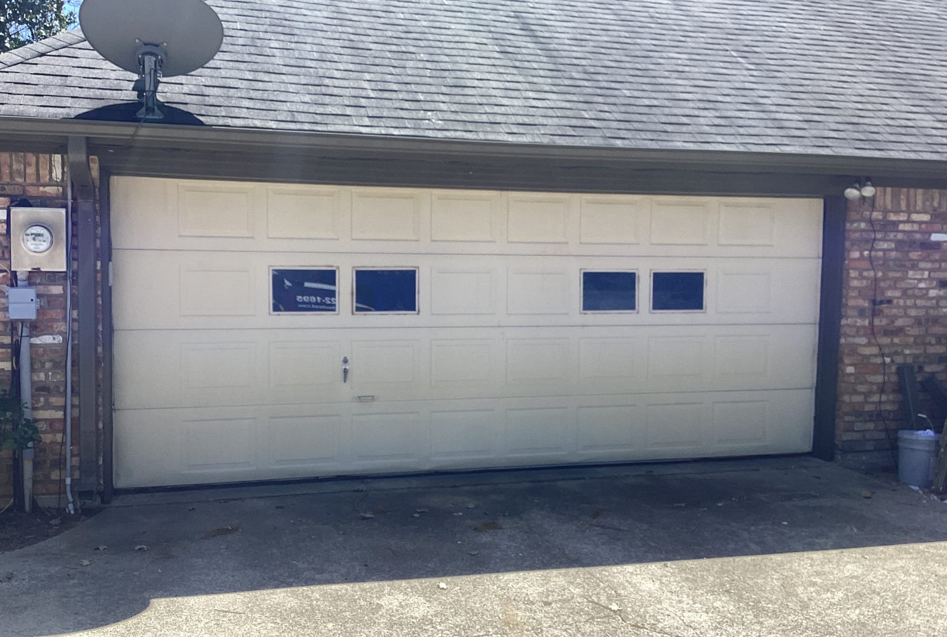 A beige garage door with two sets of windows, between brick walls, under a gray roof.