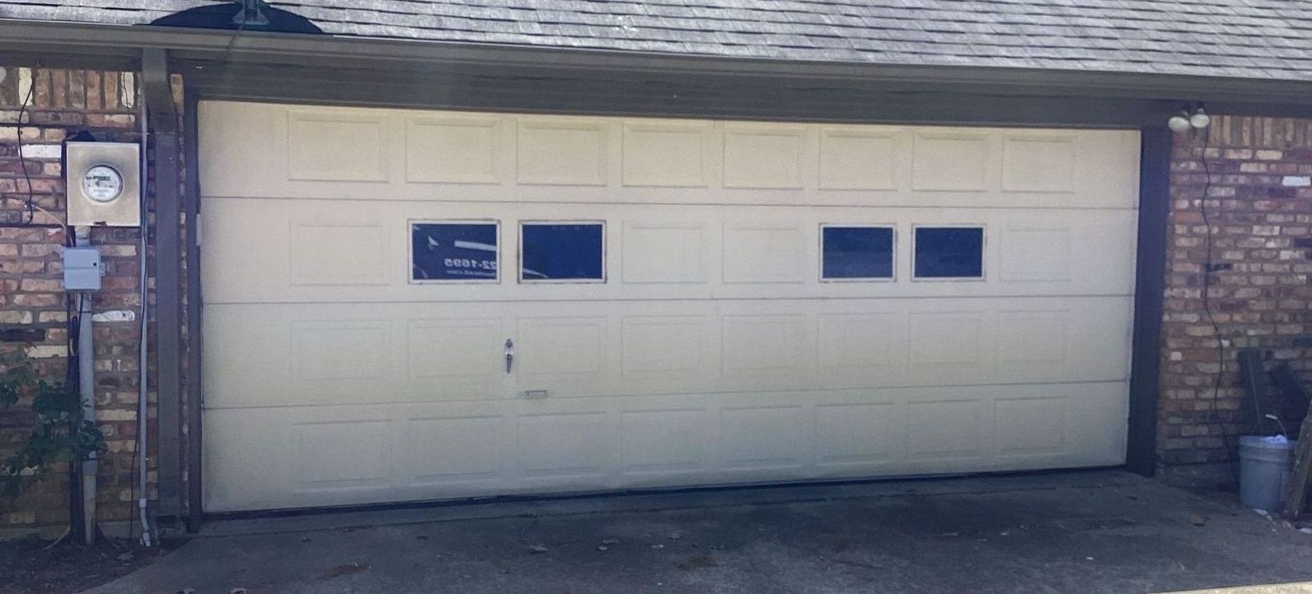 A beige garage door with two sets of windows, between brick walls, under a gray roof.