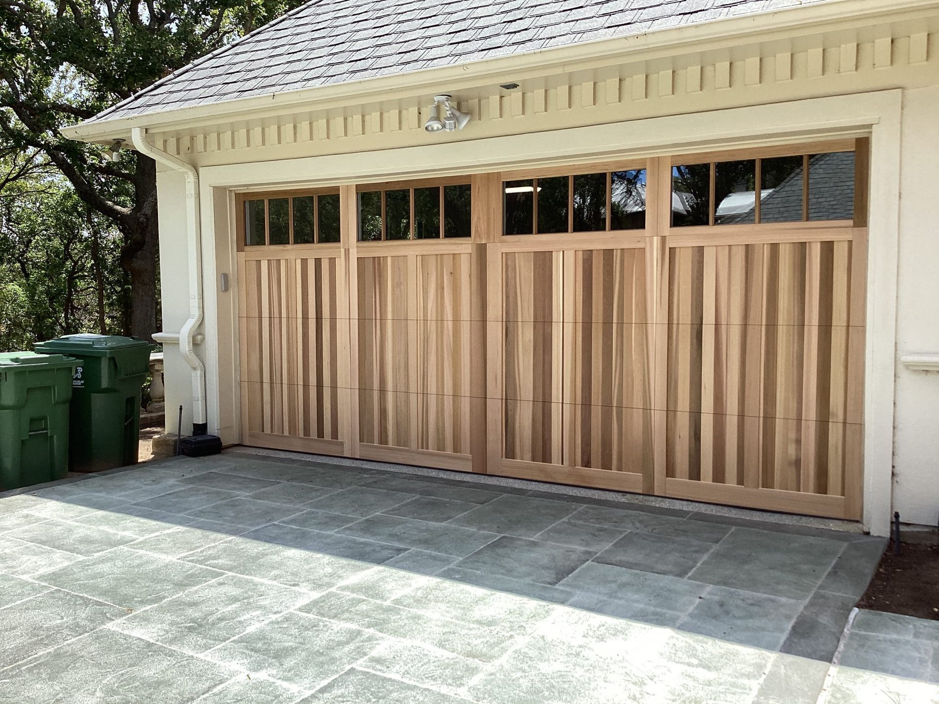 A wooden garage door with vertical slats and upper glass panels set against a stone driveway and white exterior wall.