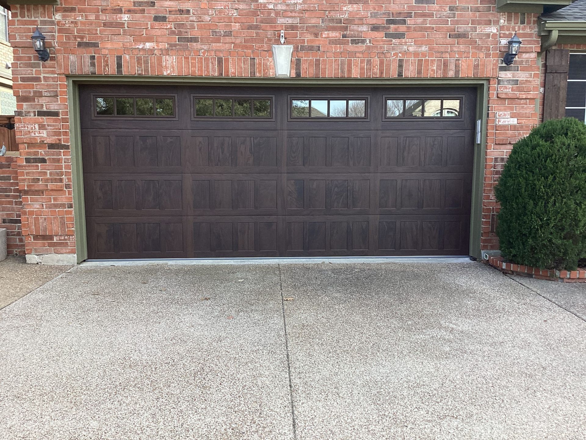 A dark brown, four-panel residential garage door with a row of windows across the top, set in a red brick exterior.