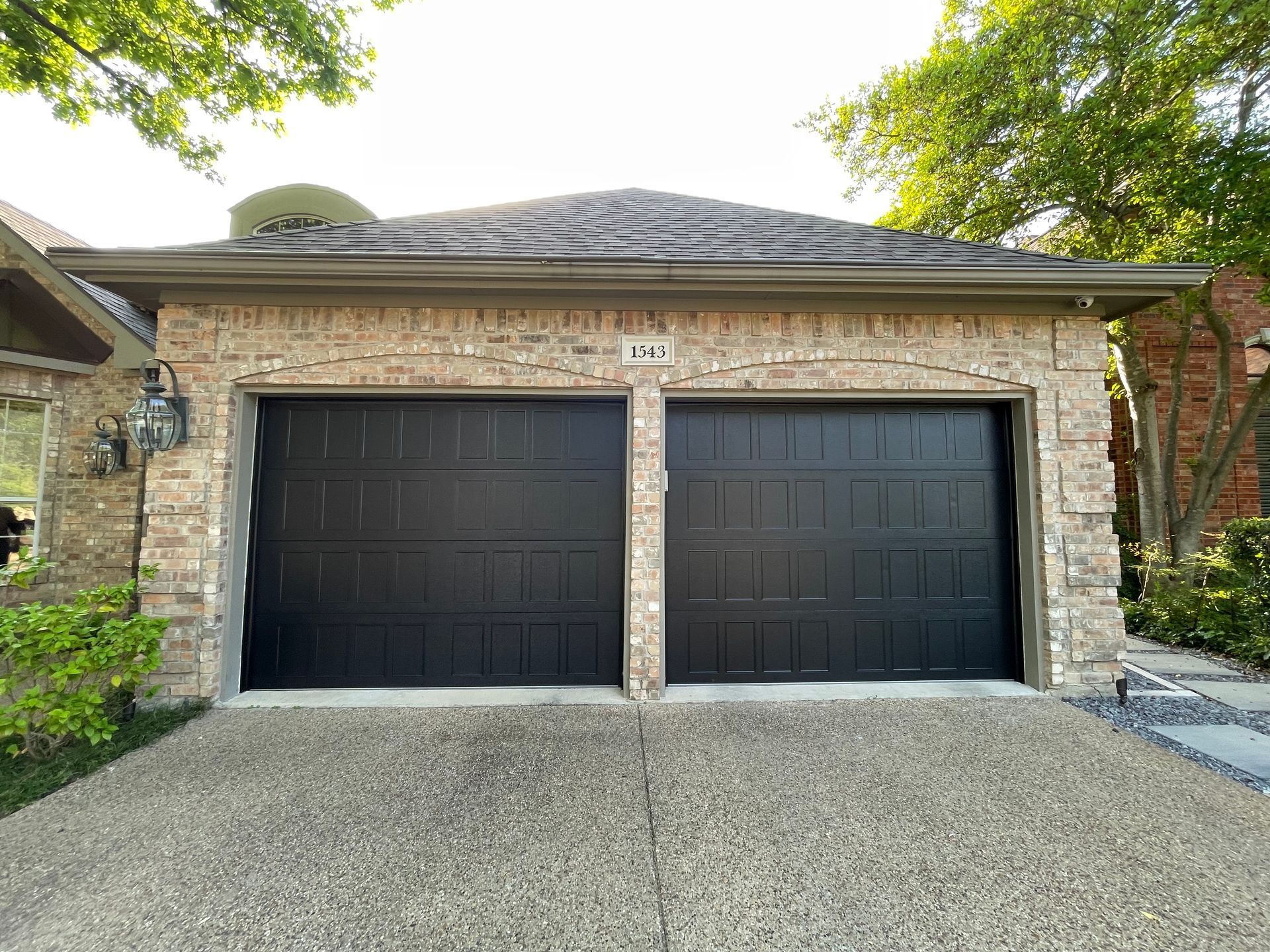 A two-car garage with black paneled doors and a light tan brick exterior, viewed from a pebble driveway.