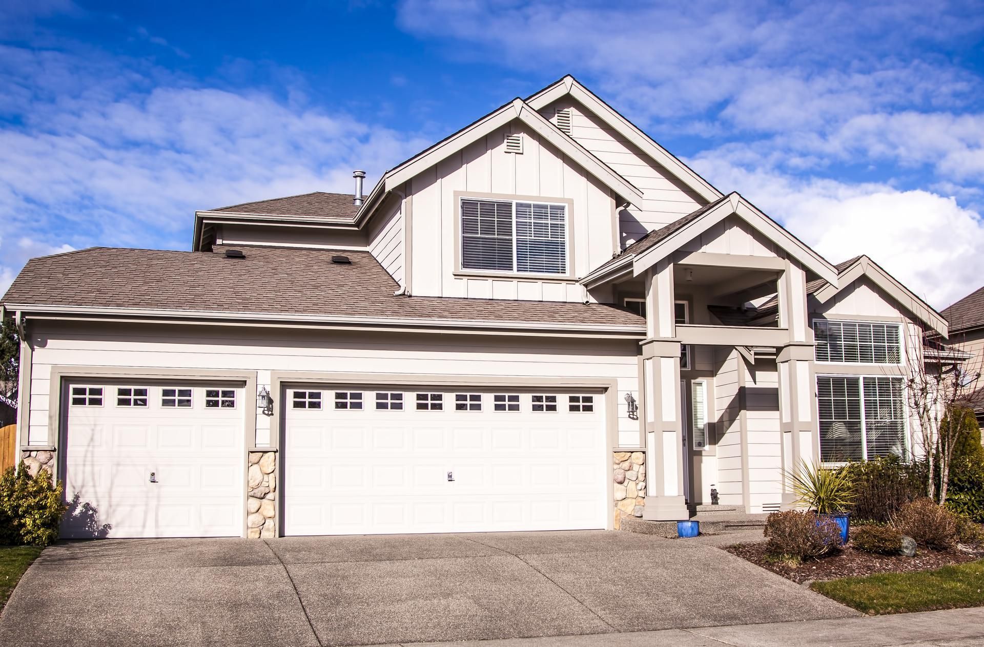 Two-story light gray house with attached garage, stone accents, and blue sky.