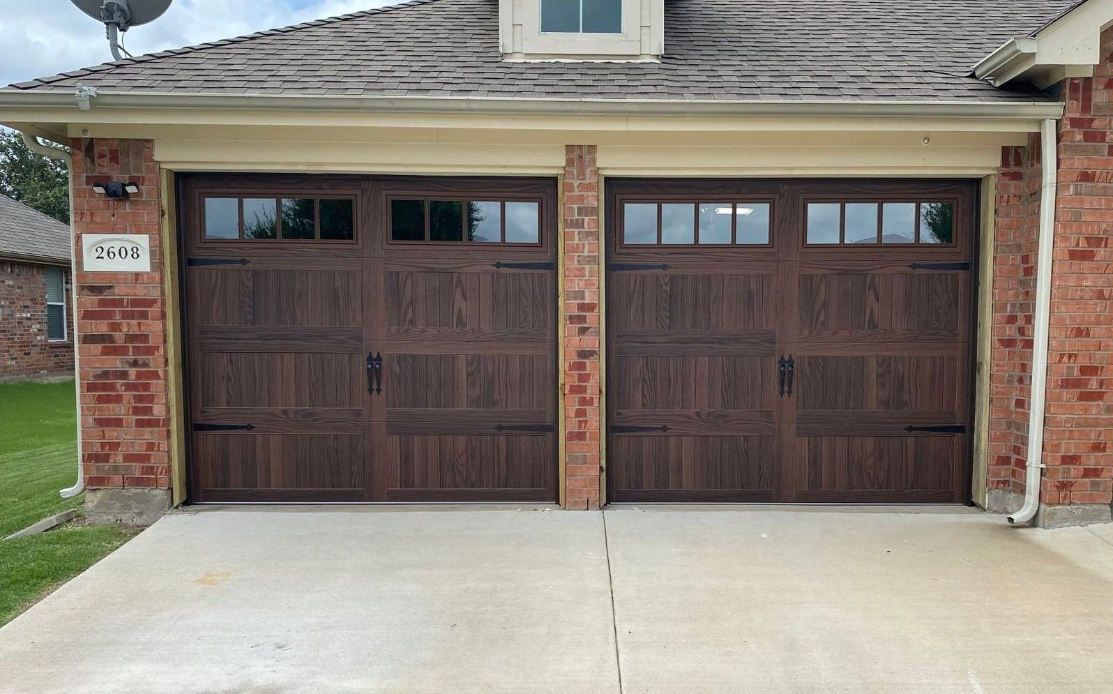 Two brown wooden garage doors with windows, in a brick house, on a concrete driveway.