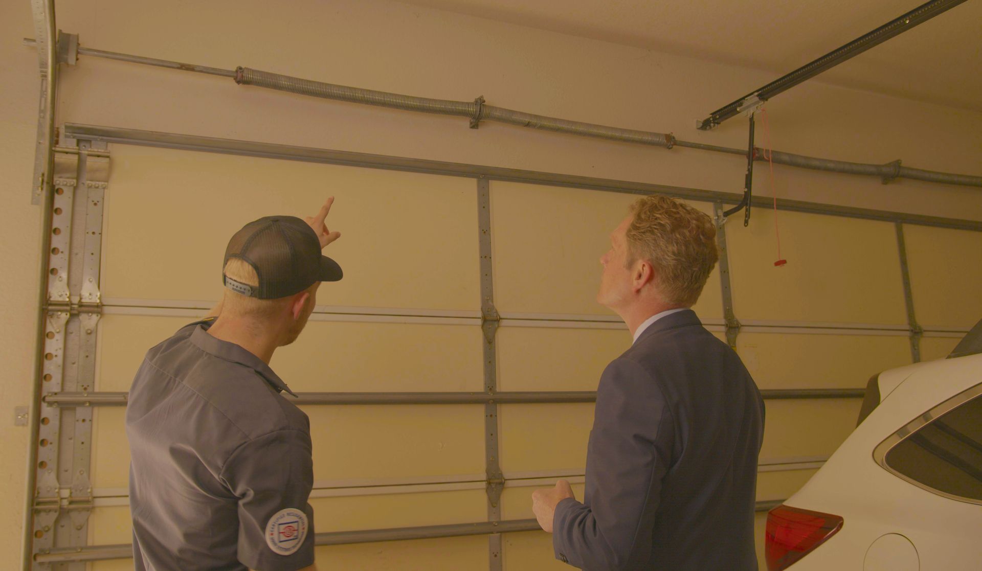 Two men examine a garage door. One points to a spring above the closed door.
