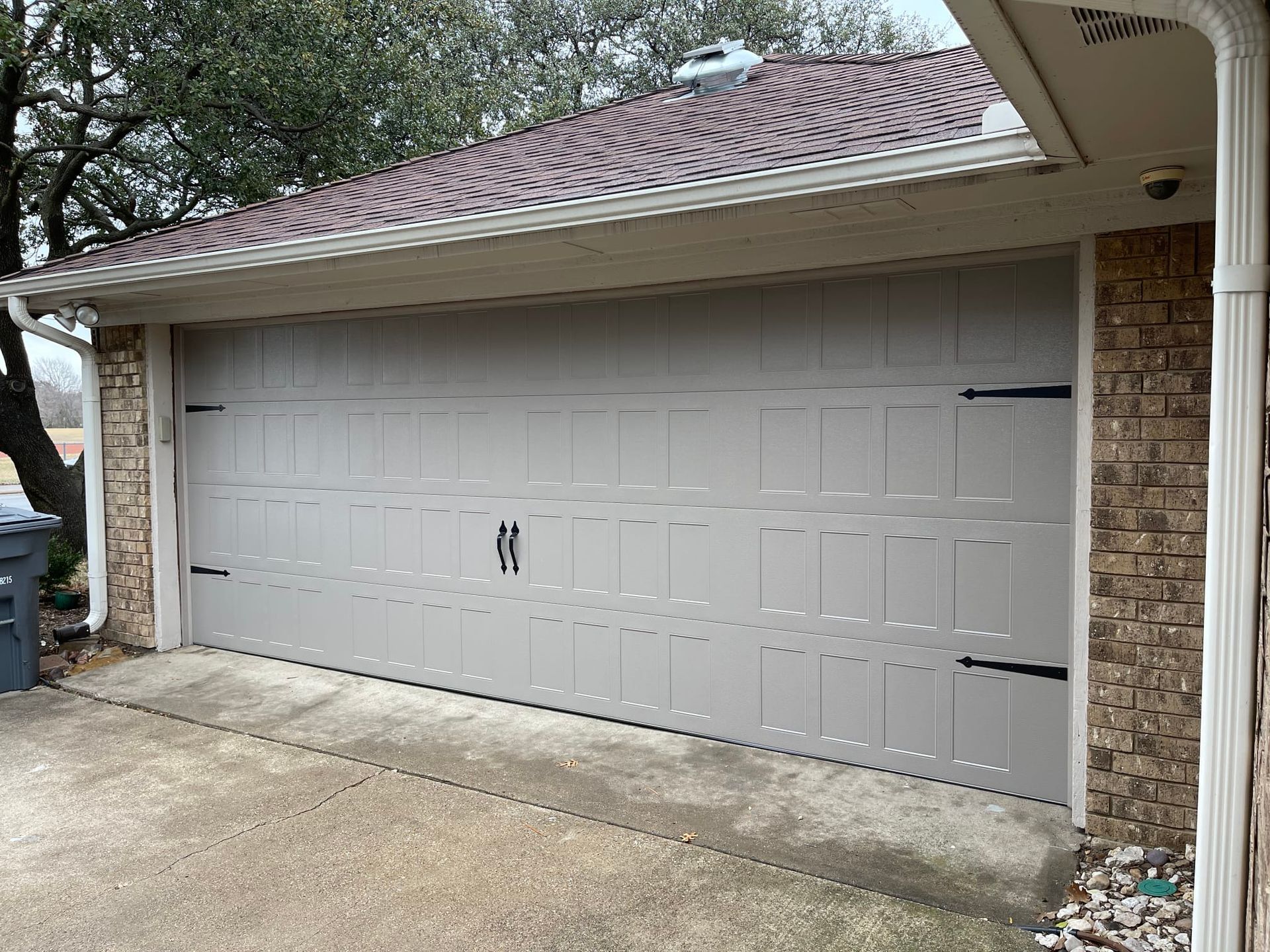 A two-car beige garage door with decorative black hinges and handles, installed in a brick house with a shingled roof.