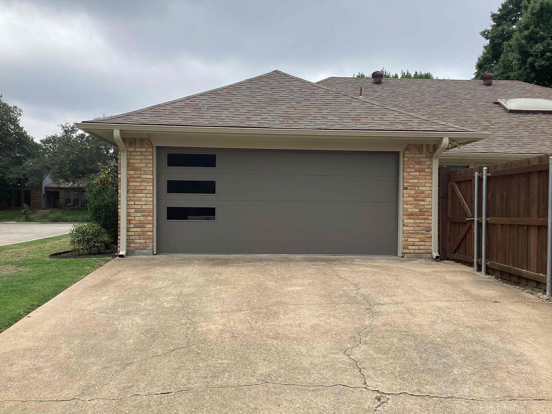 Gray garage door with three rectangular windows, brick facade, concrete driveway.