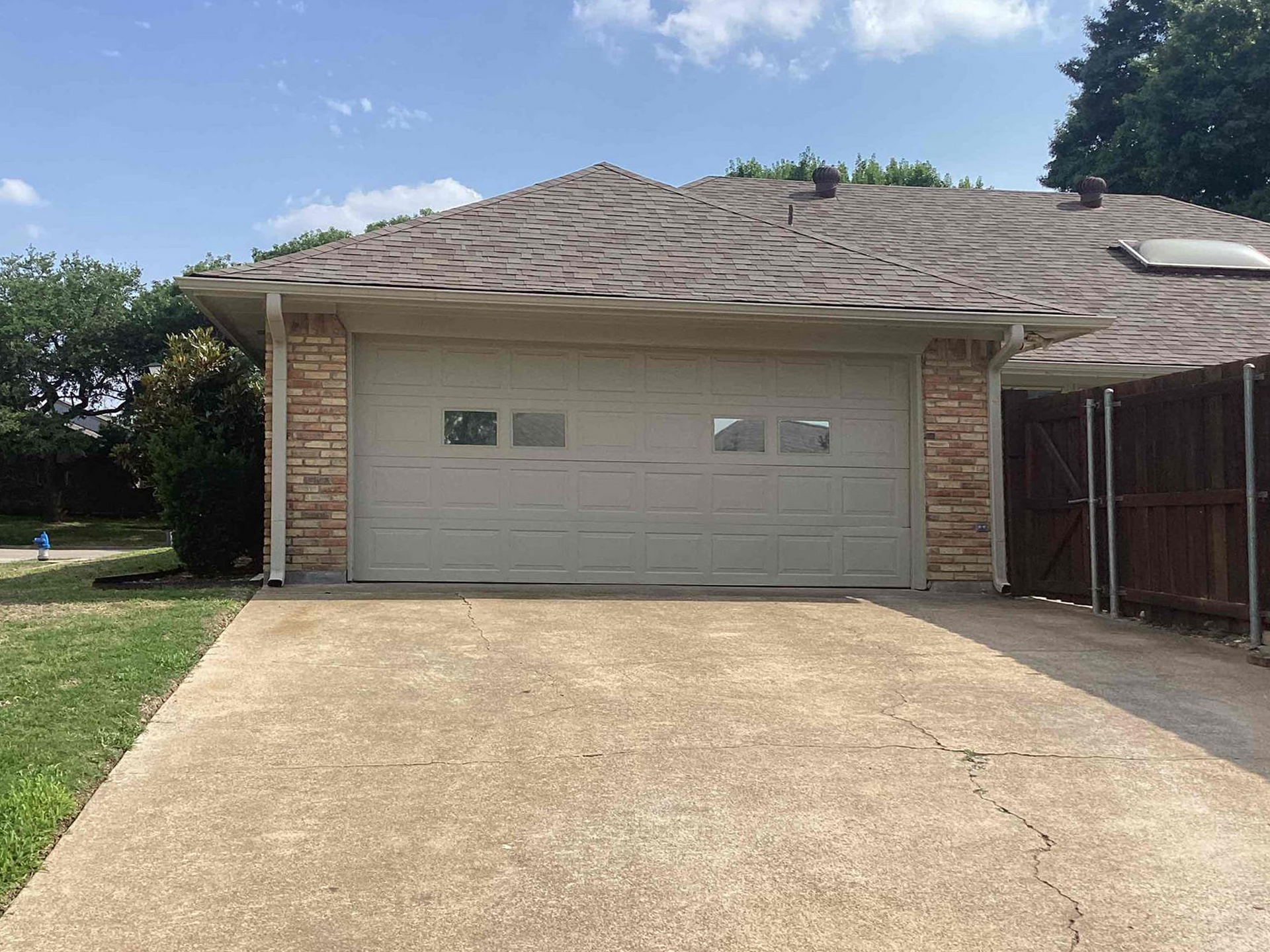 Gray garage door on a concrete driveway, beige brick siding, brown roof, and blue sky.