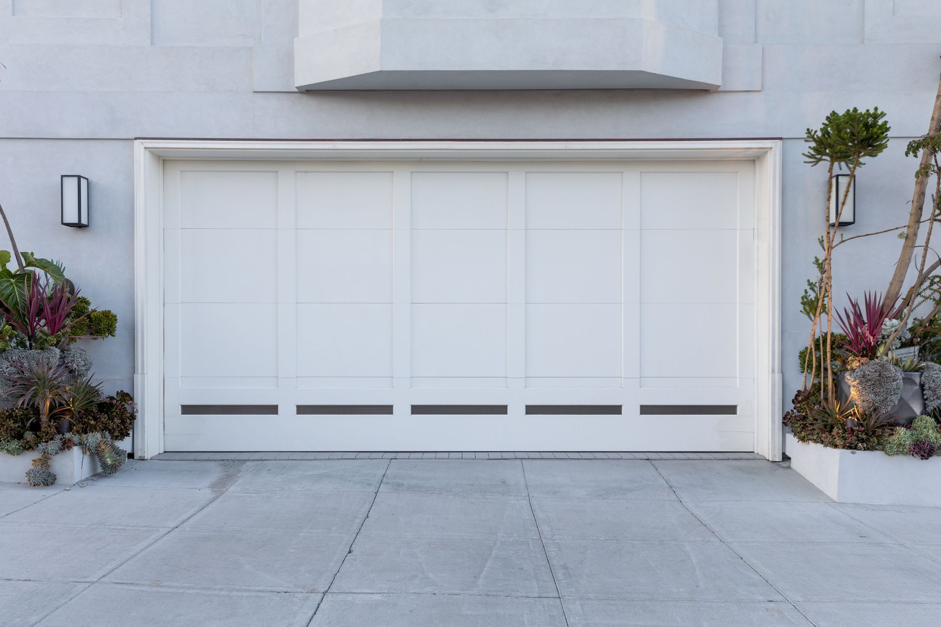 White garage door, flanked by greenery and lights, set in a gray concrete driveway.