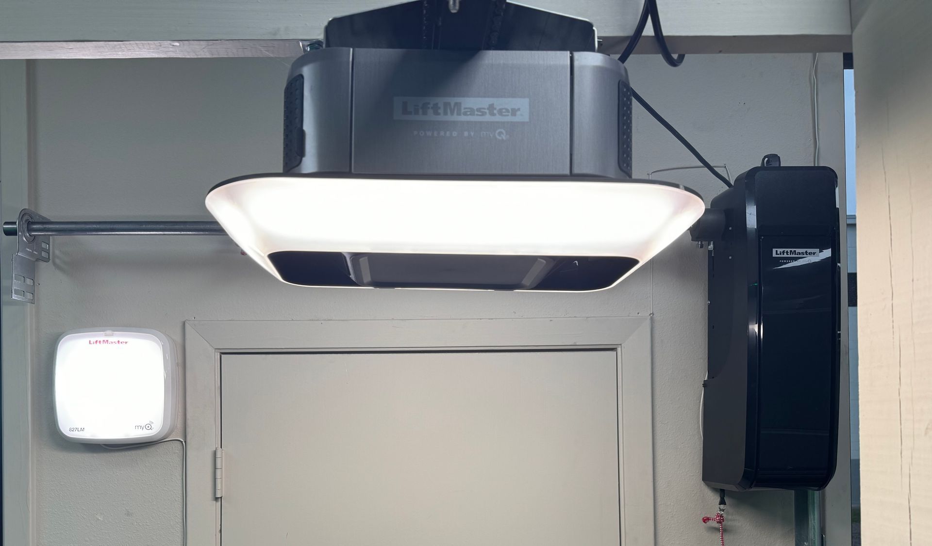 Garage door opener with light above a closed white door. A wall-mounted control and light are also visible.