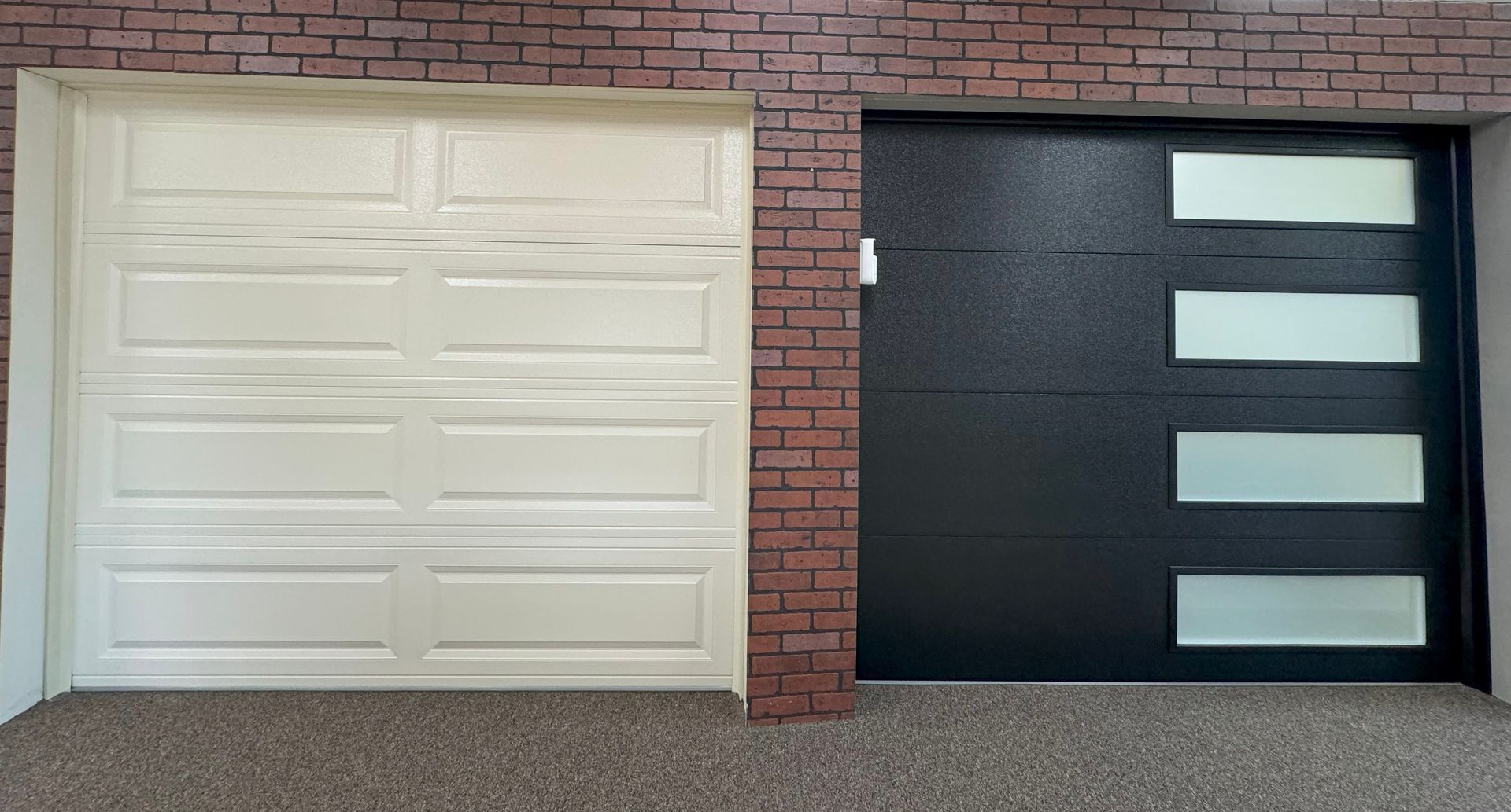 Two garage doors, one white, paneled; the other black with four glass windows. Brick wall.