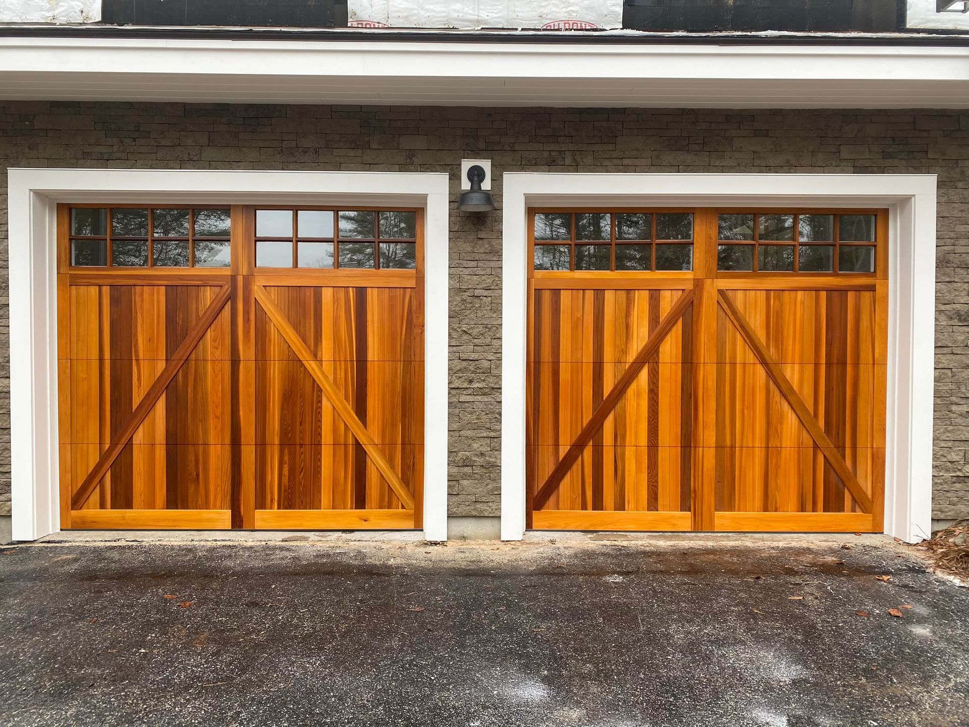 Two wooden garage doors with glass window panels and diagonal cross-bracing, set into a stone wall.