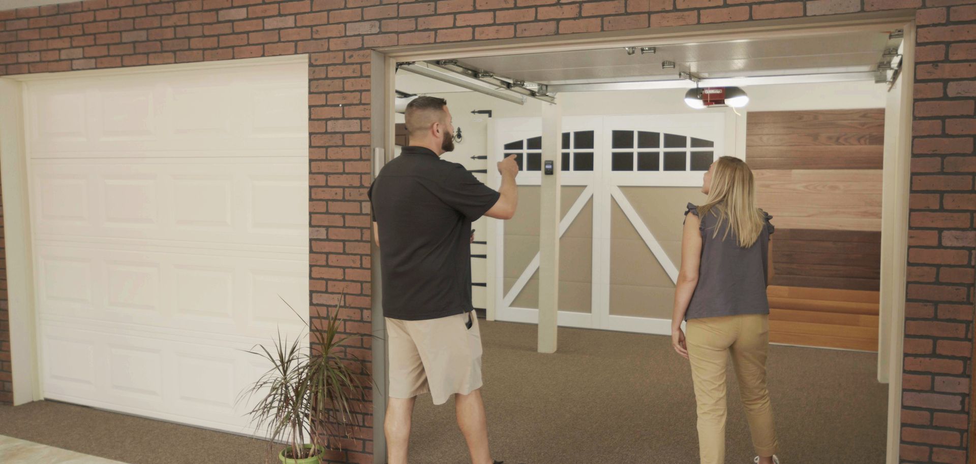 Two people walking into a garage. Man holds up a phone, woman looks ahead. Beige and white garage doors. Brick walls.