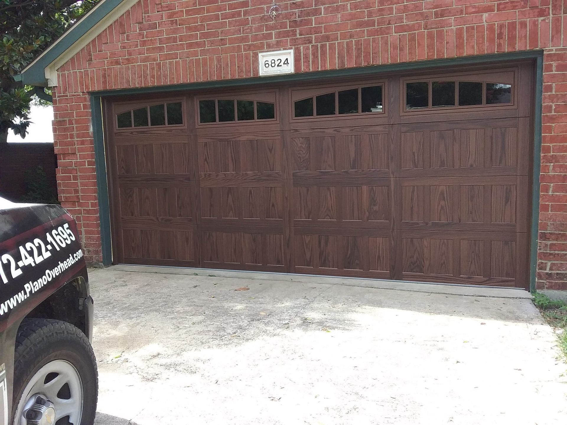 A dark wood-toned garage door with windows installed on a brick house, partially obscured by a service truck on the left.