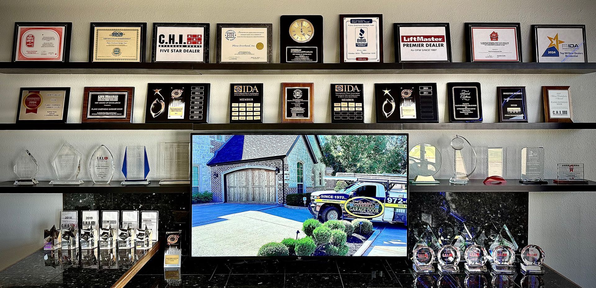 A wall with framed awards above a counter with trophies and a TV displaying a building and a vehicle.