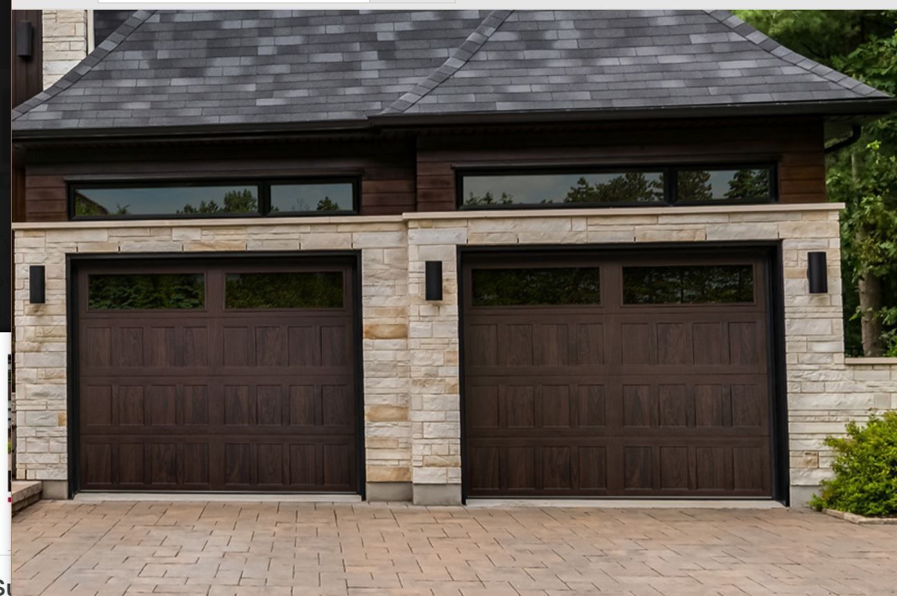 Two dark brown garage doors with top windows set in a light-colored stone wall under a dark shingled roof.