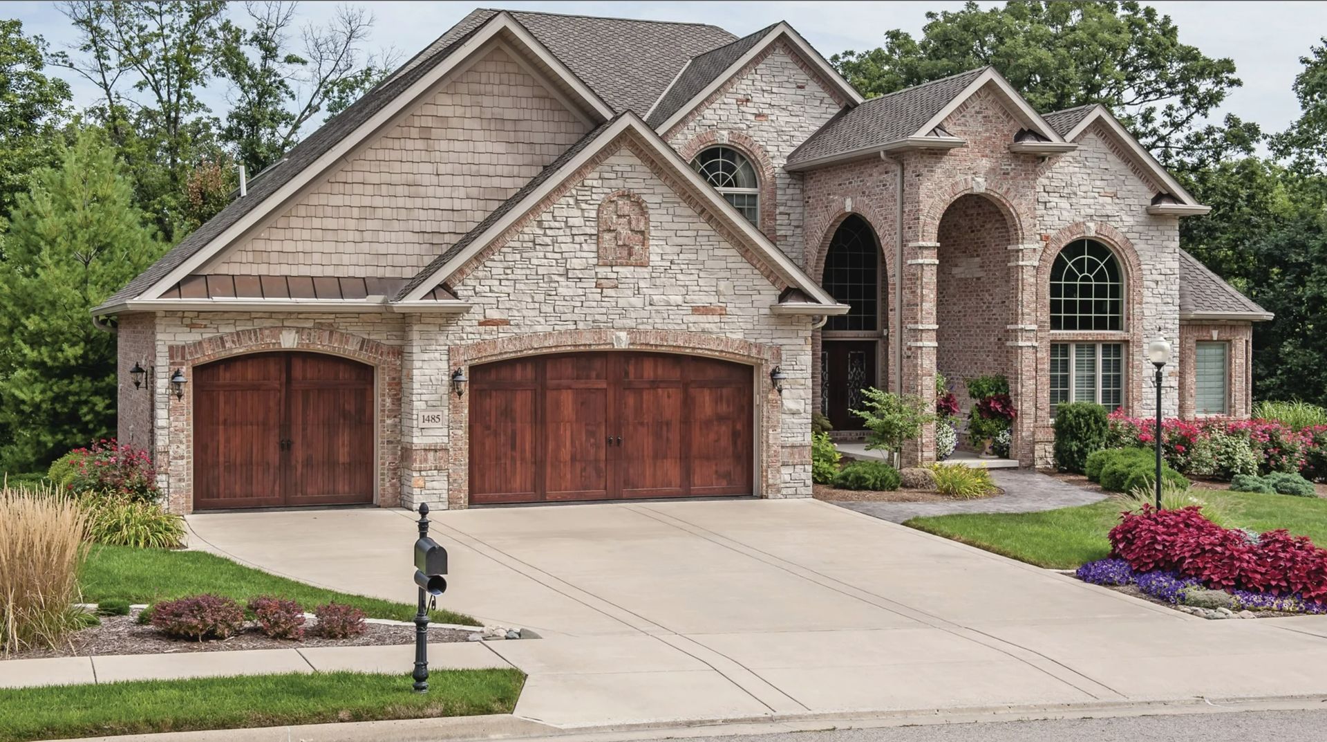 A two-story stone house with a three-car wooden garage, surrounded by a lawn and landscaping on a sunny day.