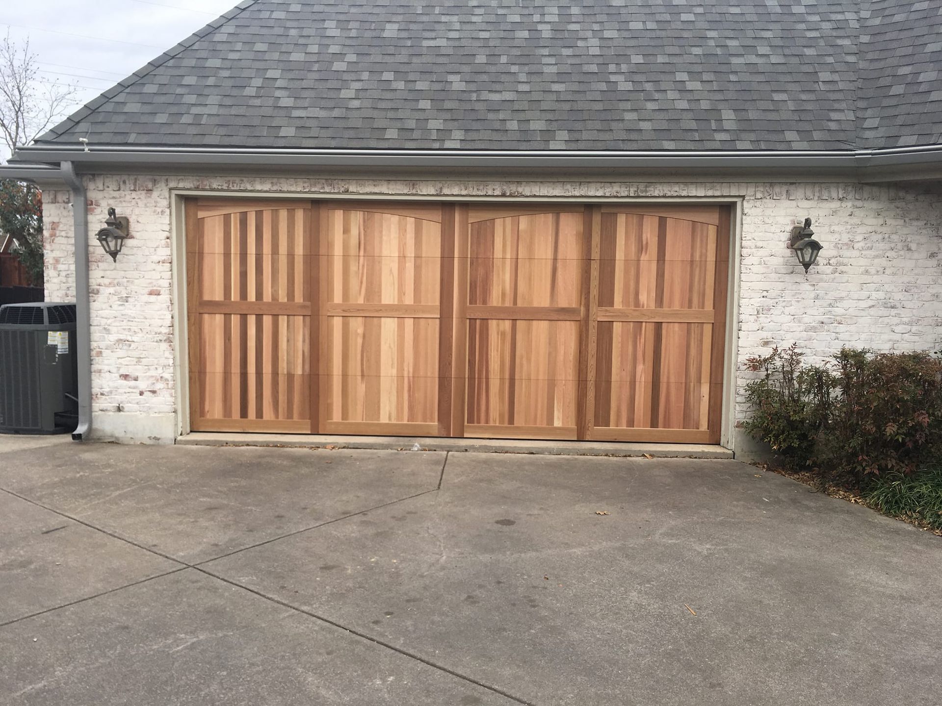 A four-panel wooden garage door with vertical slats, set within a rustic white brick house wall above a concrete driveway.
