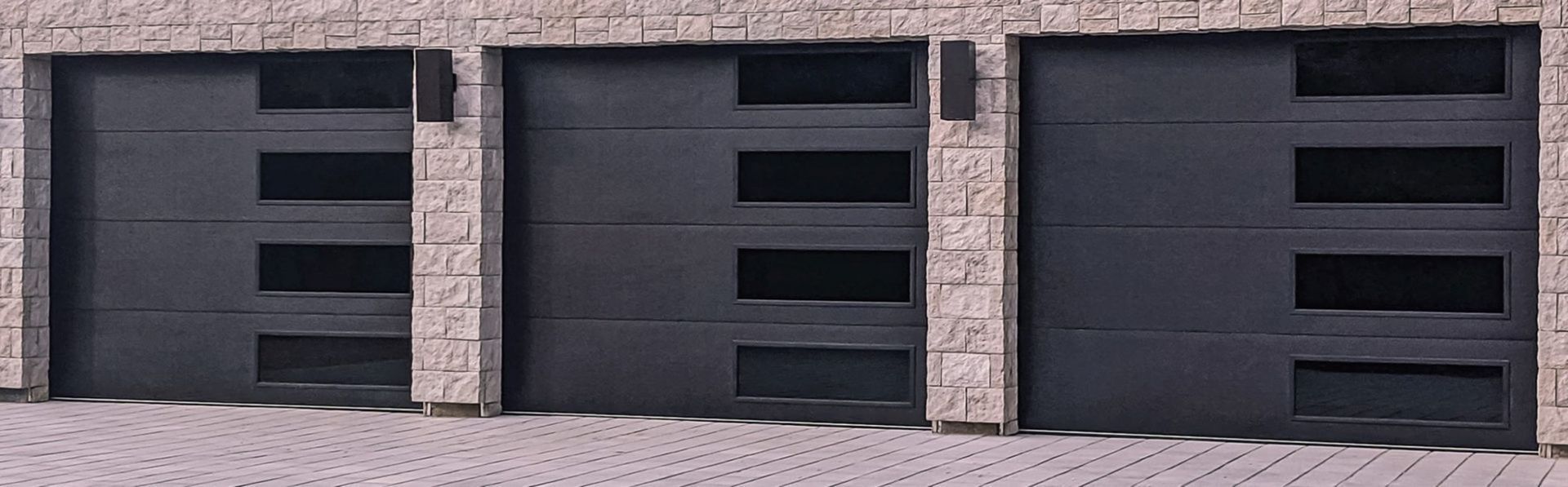 Three dark gray garage doors with rectangular windows, brick facade, paving stones.