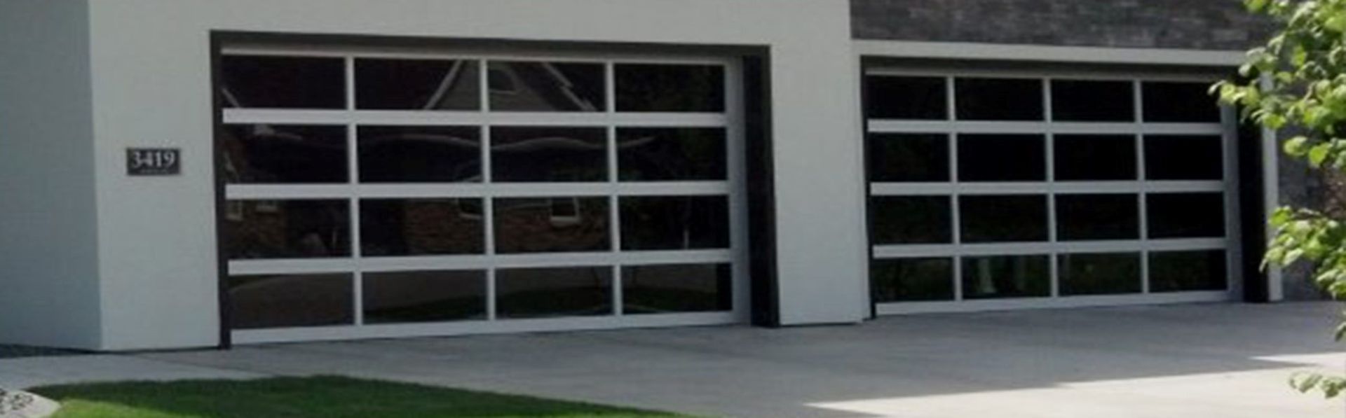 Two modern garage doors with glass panels and white frames. Concrete driveway and green lawn.